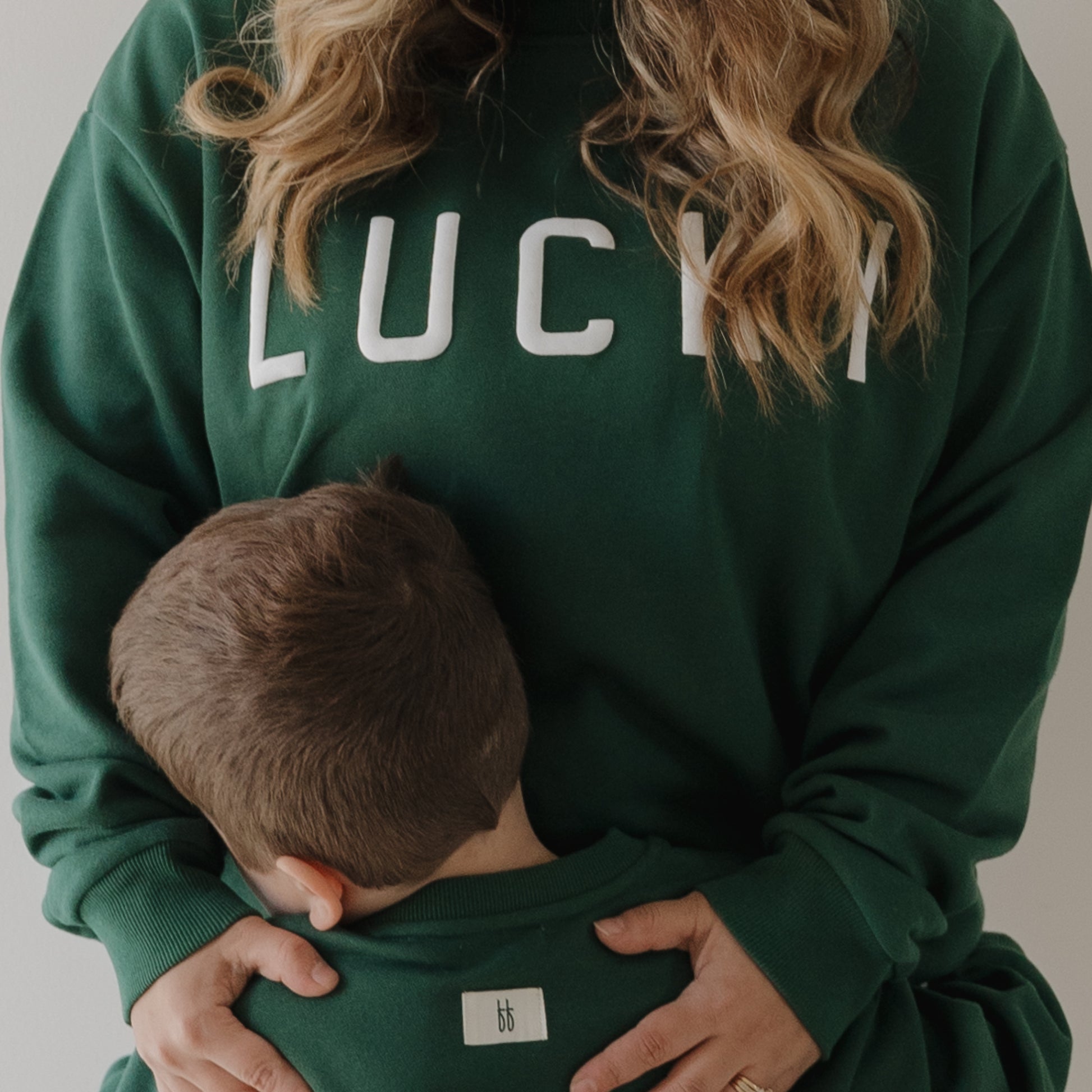 A woman wears the forever french baby Adult Sweatshirt | Lucky in green as she hugs a young boy, both dressed in matching green tops—capturing warmth and comfort, perfect inspiration for family sweatshirts.