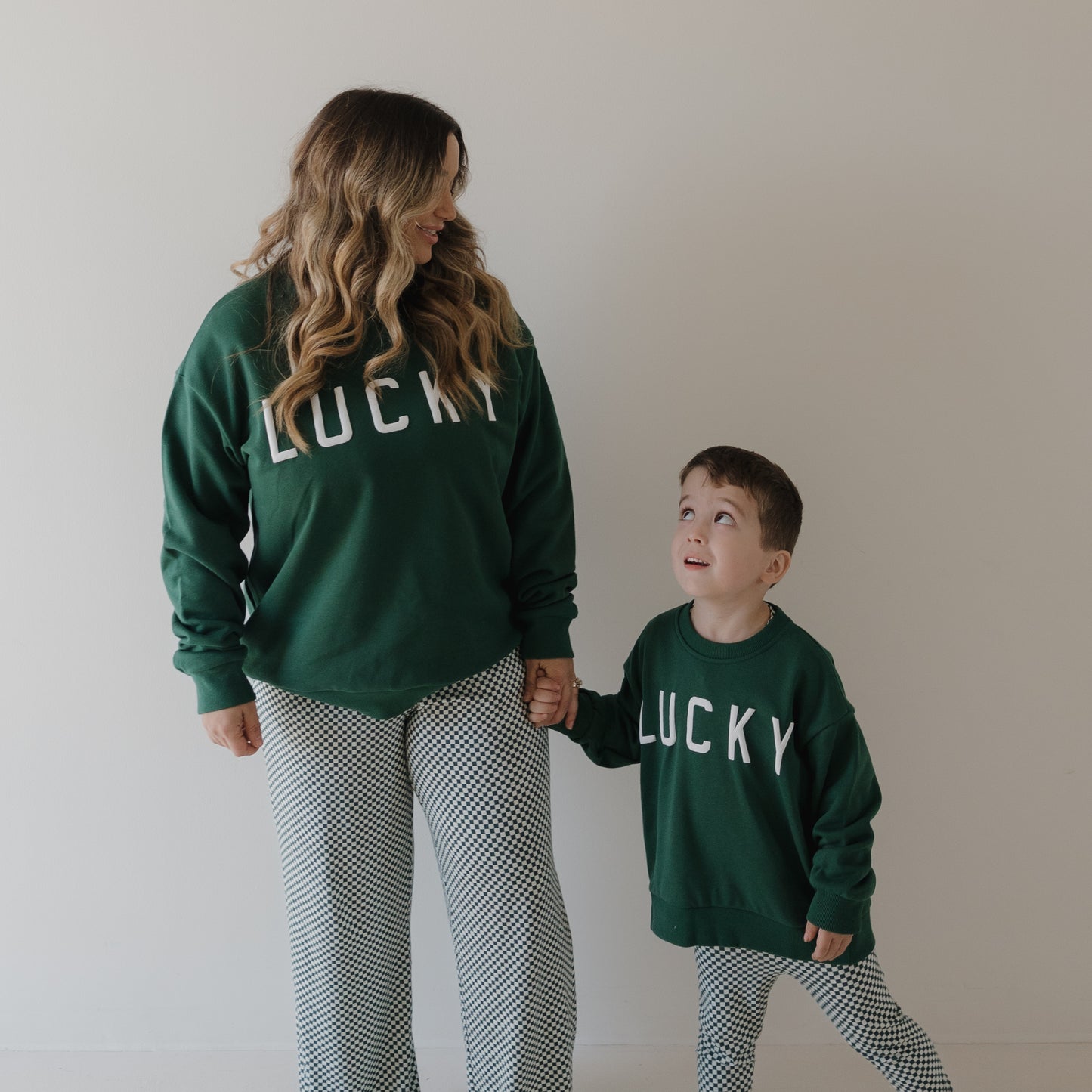 A woman and young boy smile at each other, holding hands in matching forever french baby "Lucky" adult sweatshirts and checkered pants, set against a plain background—perfect for your family St. Patrick’s Day outfit.