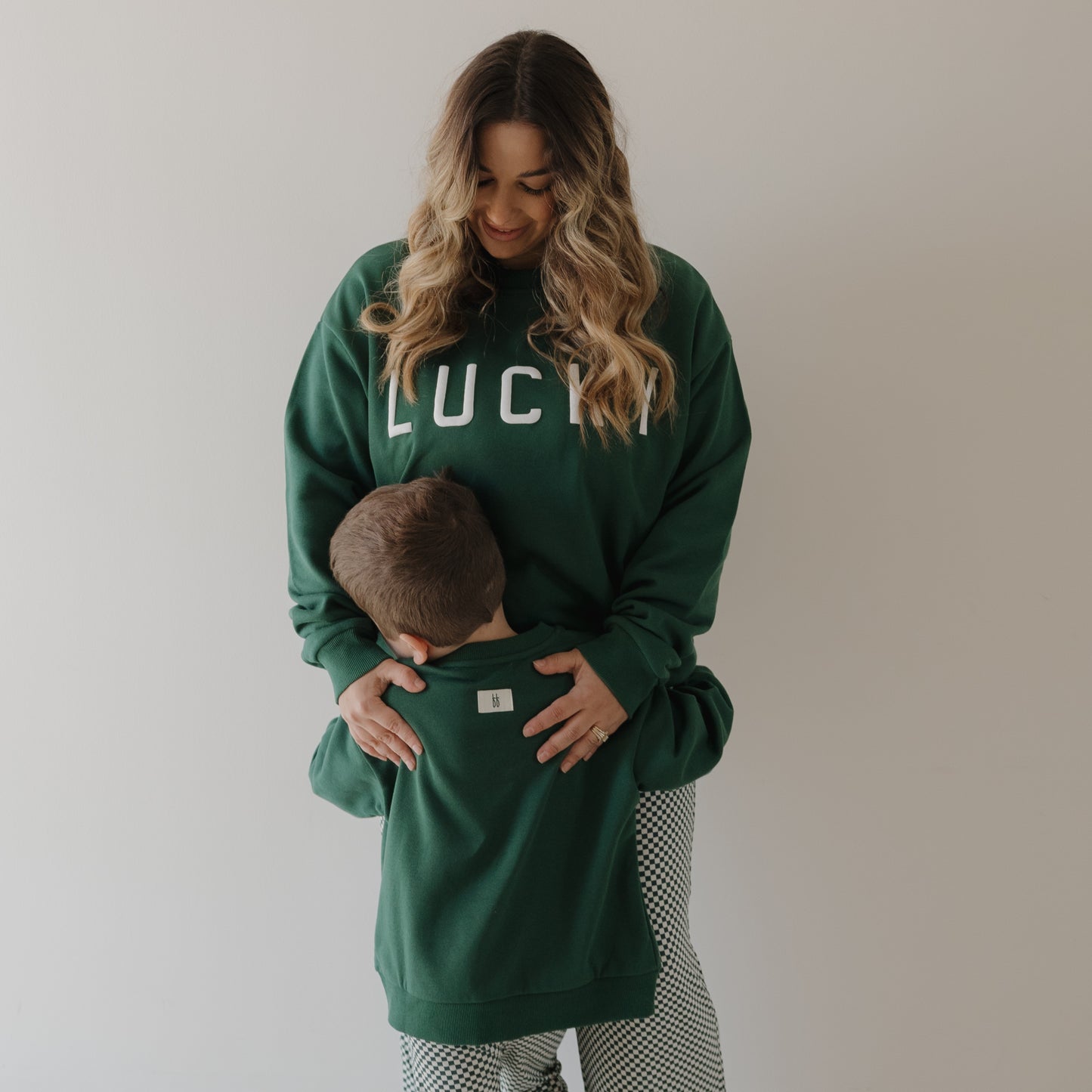A woman in a green forever french baby "Lucky" Adult Sweatshirt smiles at a young boy hugging her waist. Both wear matching family sweatshirts, posing casually against a plain, light background.