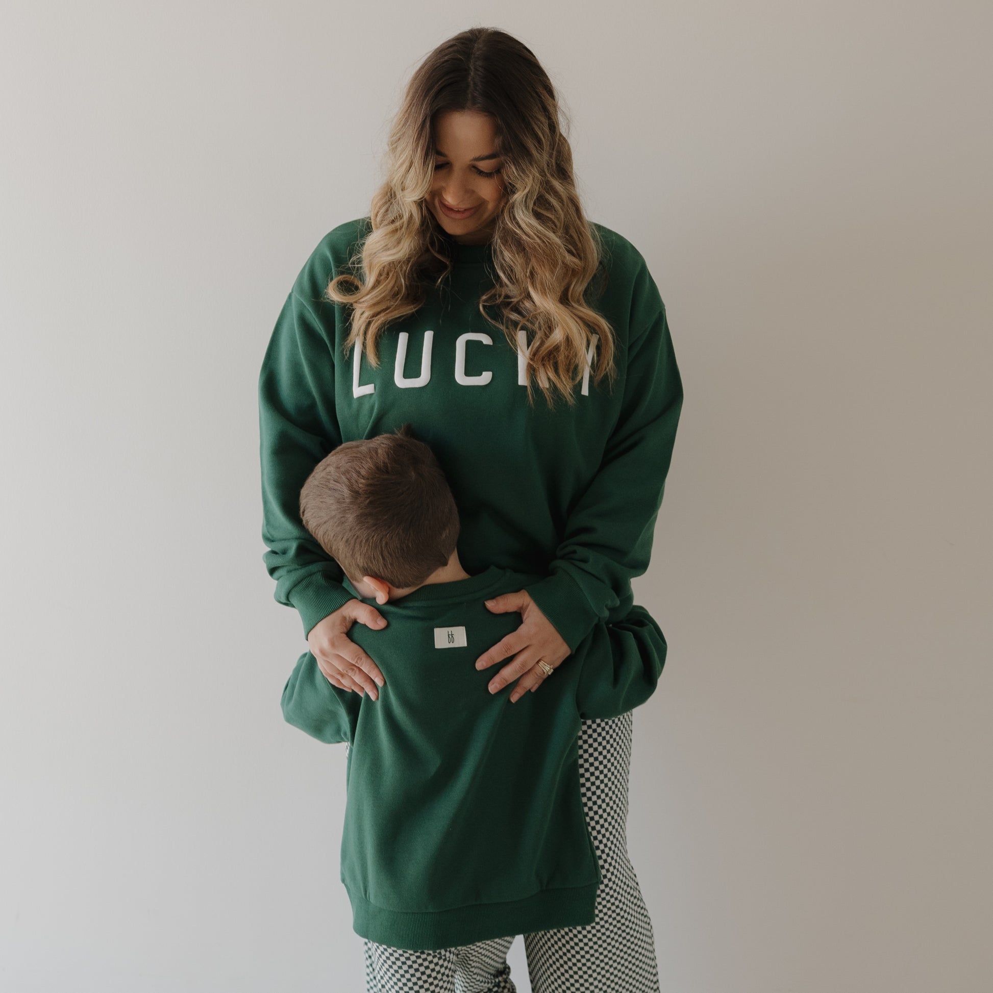 A woman in a green forever french baby "Lucky" Adult Sweatshirt smiles at a young boy hugging her waist. Both wear matching family sweatshirts, posing casually against a plain, light background.