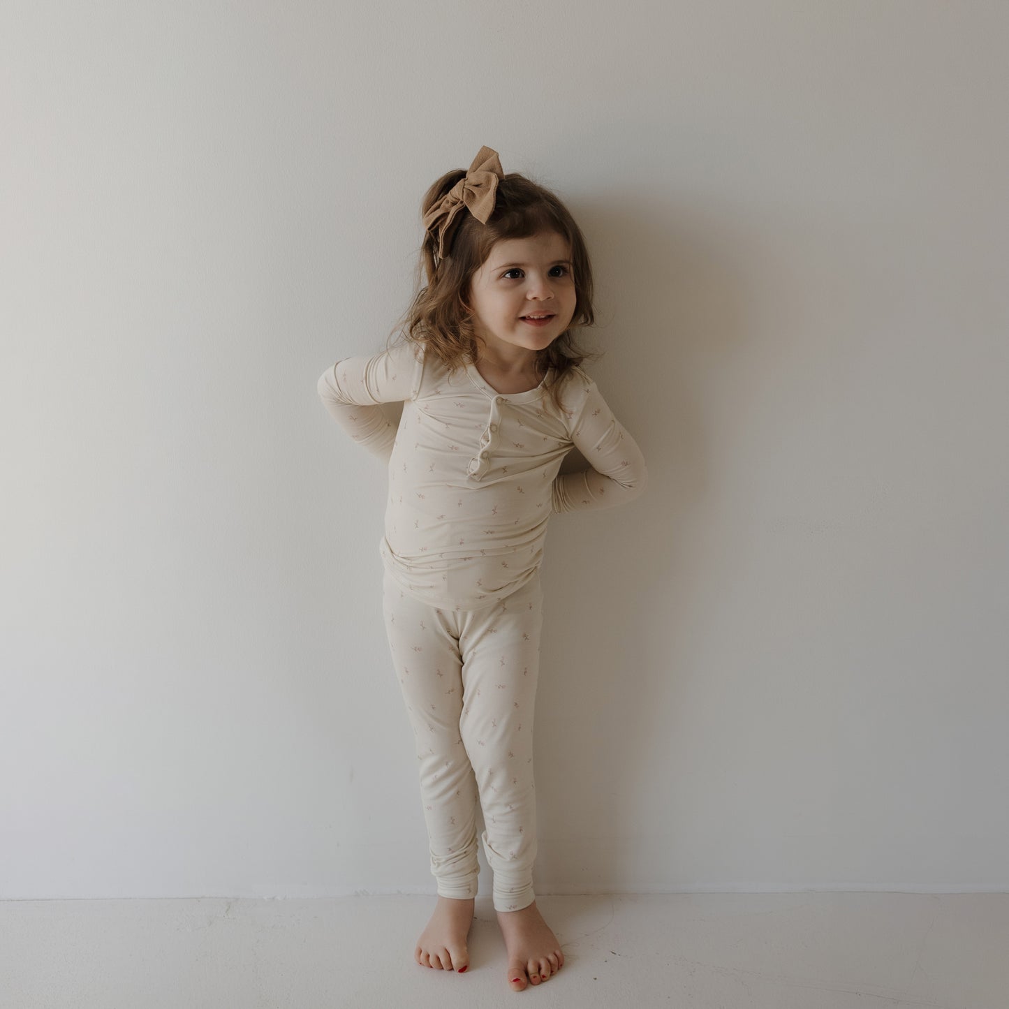 A young girl with brown hair wears forever french baby’s Bamboo Two Piece Pajamas | Lucy and a large tan bow, standing barefoot against a white wall and smiling with her hands behind her back.