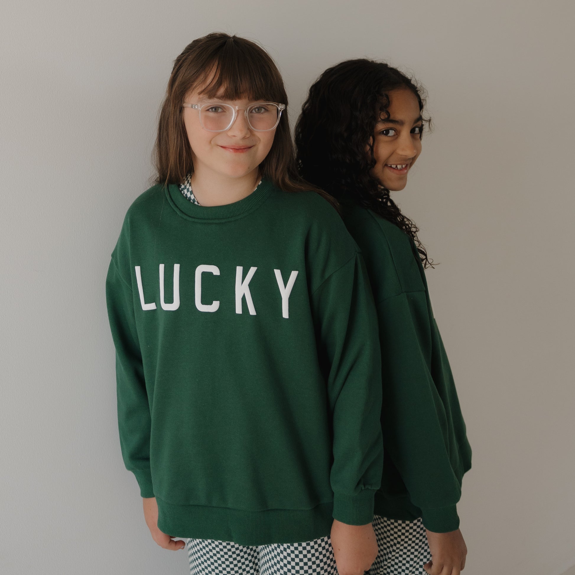 Two girls stand back-to-back in forever french baby Child Sweatshirt | Lucky—one wearing the green "LUCKY" print for St. Patrick's Day with checkered pants. One has glasses and straight hair; the other has curly hair and a slight smile.