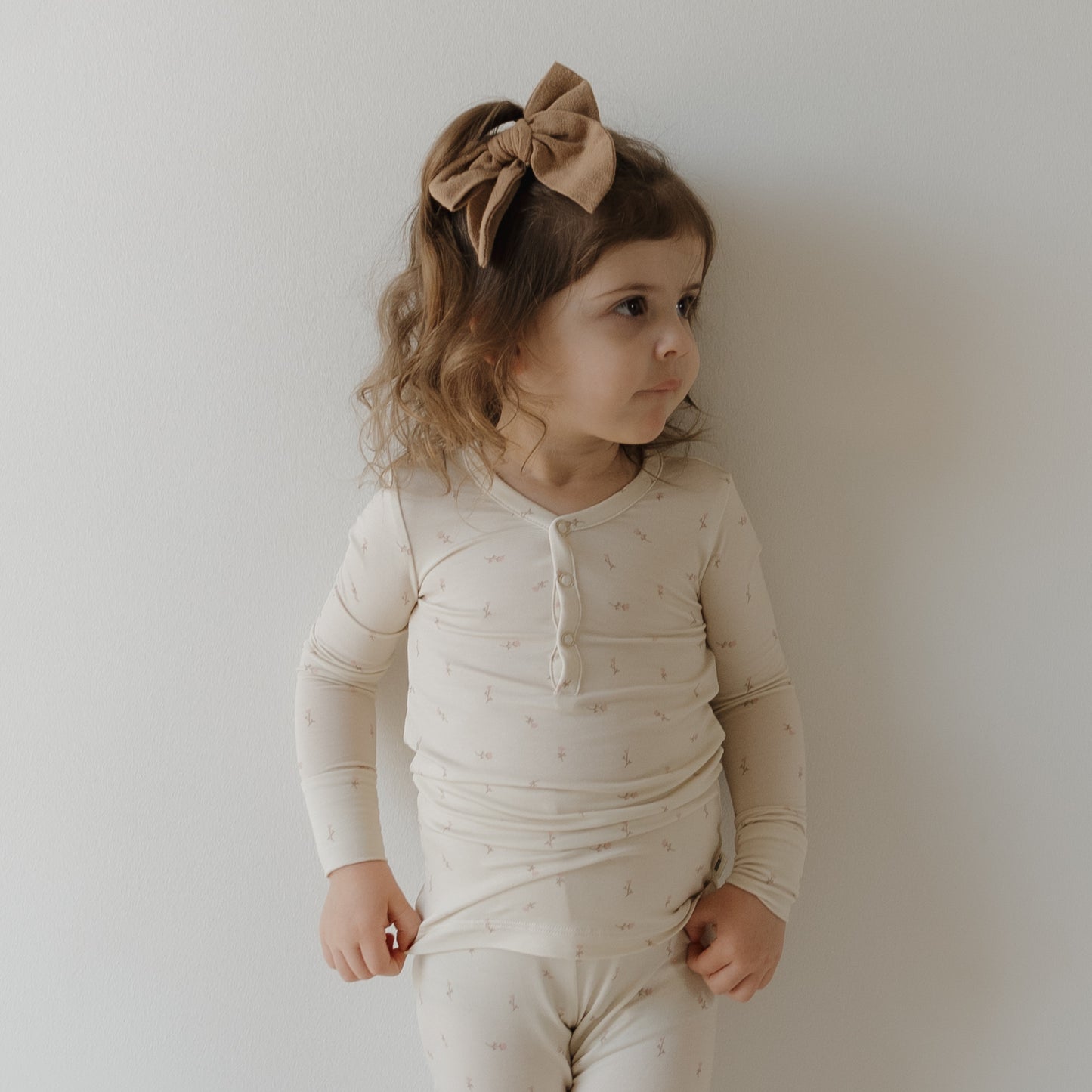 A young girl with wavy brown hair and a large tan bow wears the forever french baby Bamboo Two Piece Pajamas | Lucy, a cream-colored set with subtle patterns and matching pants, as she stands thoughtfully against a plain white wall.