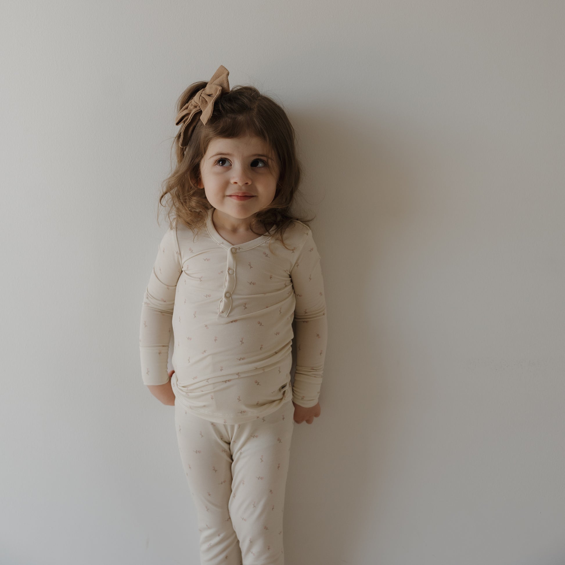 A young girl with long brown hair wears forever french baby's Bamboo Two Piece Pajamas | Lucy and a matching bow, standing against a white wall with a playful expression.