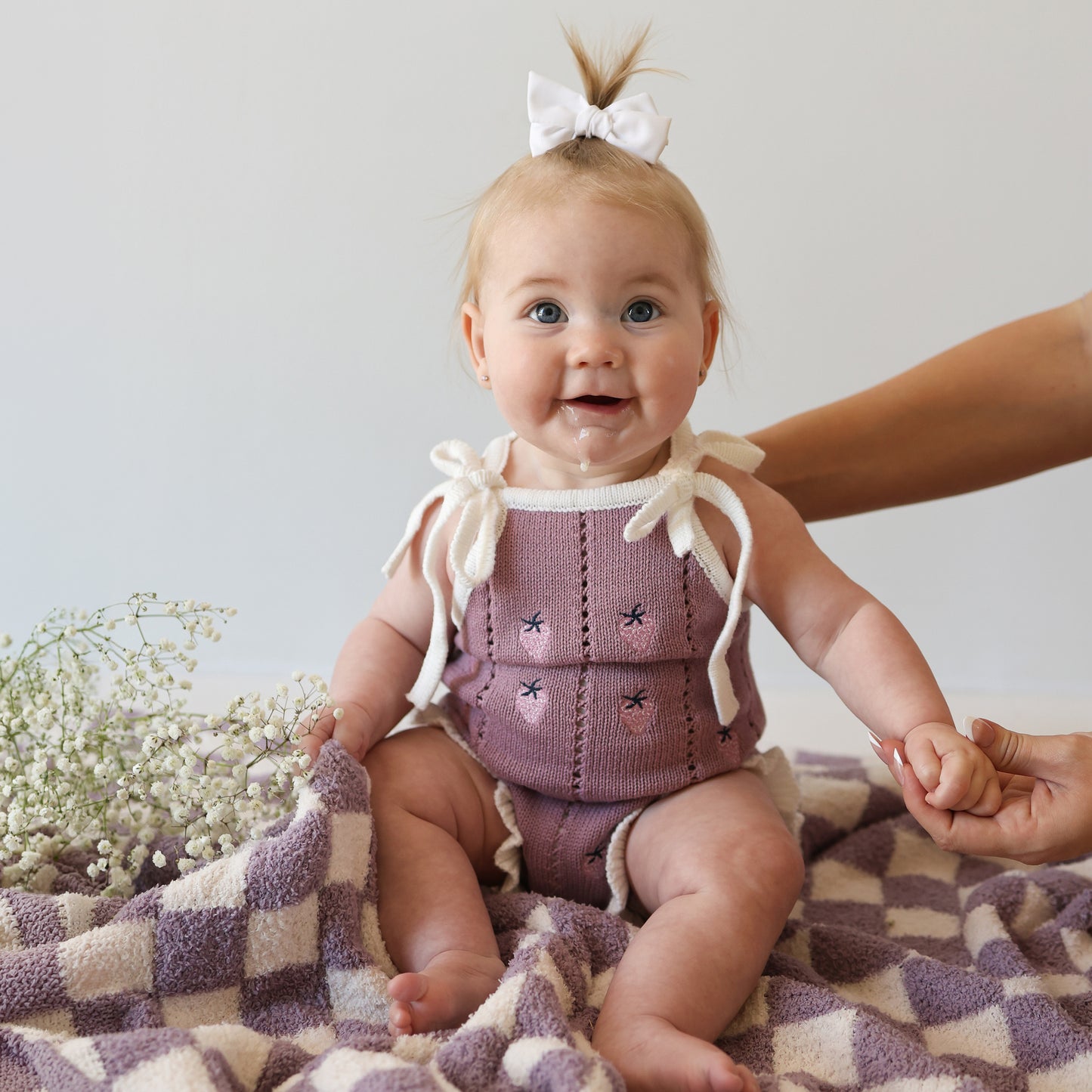 A smiling baby with a white bow sits on a checkered blanket, wearing the forever french baby Knit Ruffle Romper | Matilda with adjustable tie straps and snaps. An adult’s hand holds her arm, and white flowers are nearby.