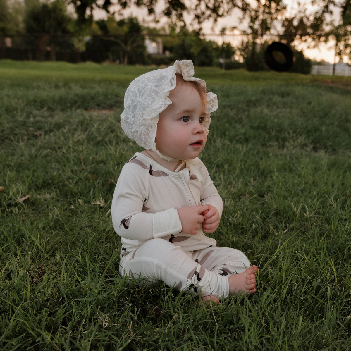 A baby in forever french baby’s Bamboo Zip Pajamas | Mallard—featuring a cream base with brown patterns—sits barefoot on a patch of green grass, gazing to the side beneath a white bonnet. The soft blur of trees and a tire swing in the background enhances the calm atmosphere, making this an ideal setting to showcase hypoallergenic babywear.