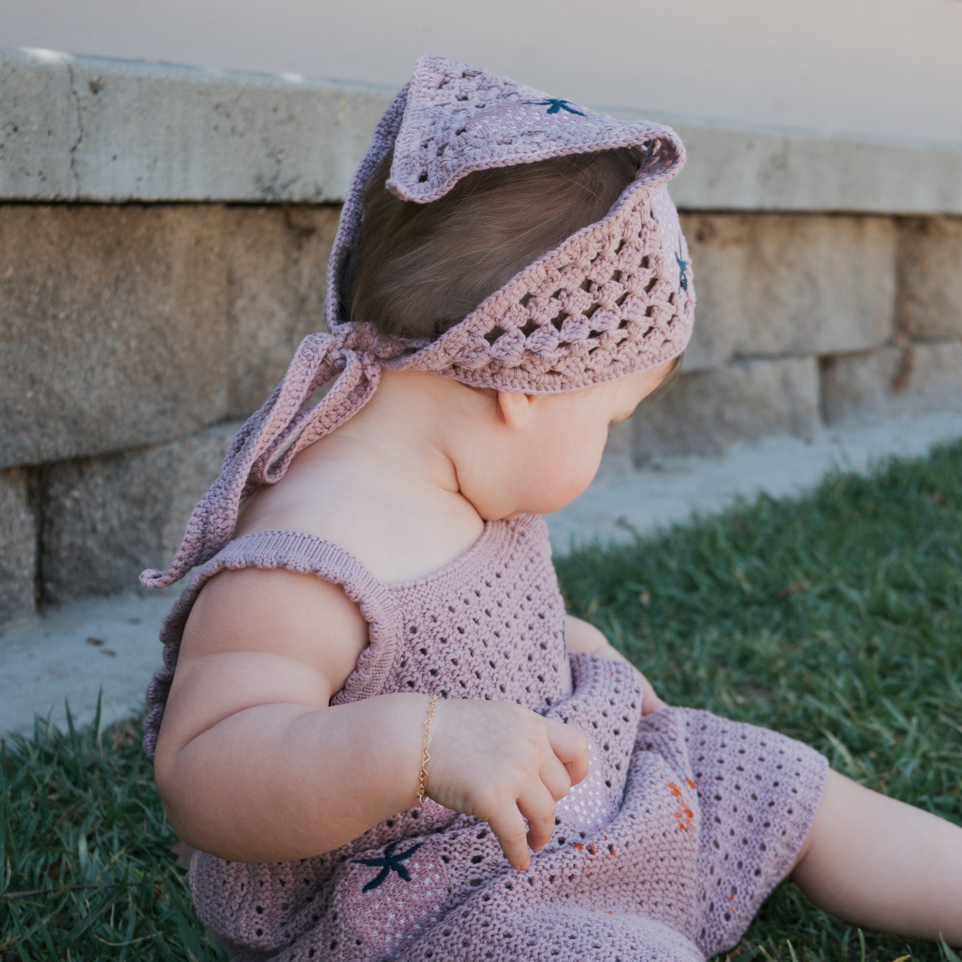 A baby wearing the forever french baby Girl's Knit Short Set | Matilda—featuring a crocheted purple tank, shorts, and a matching headscarf made from 100% cotton soft knit fabric—sits on green grass looking downward, with a stone wall in the background.
