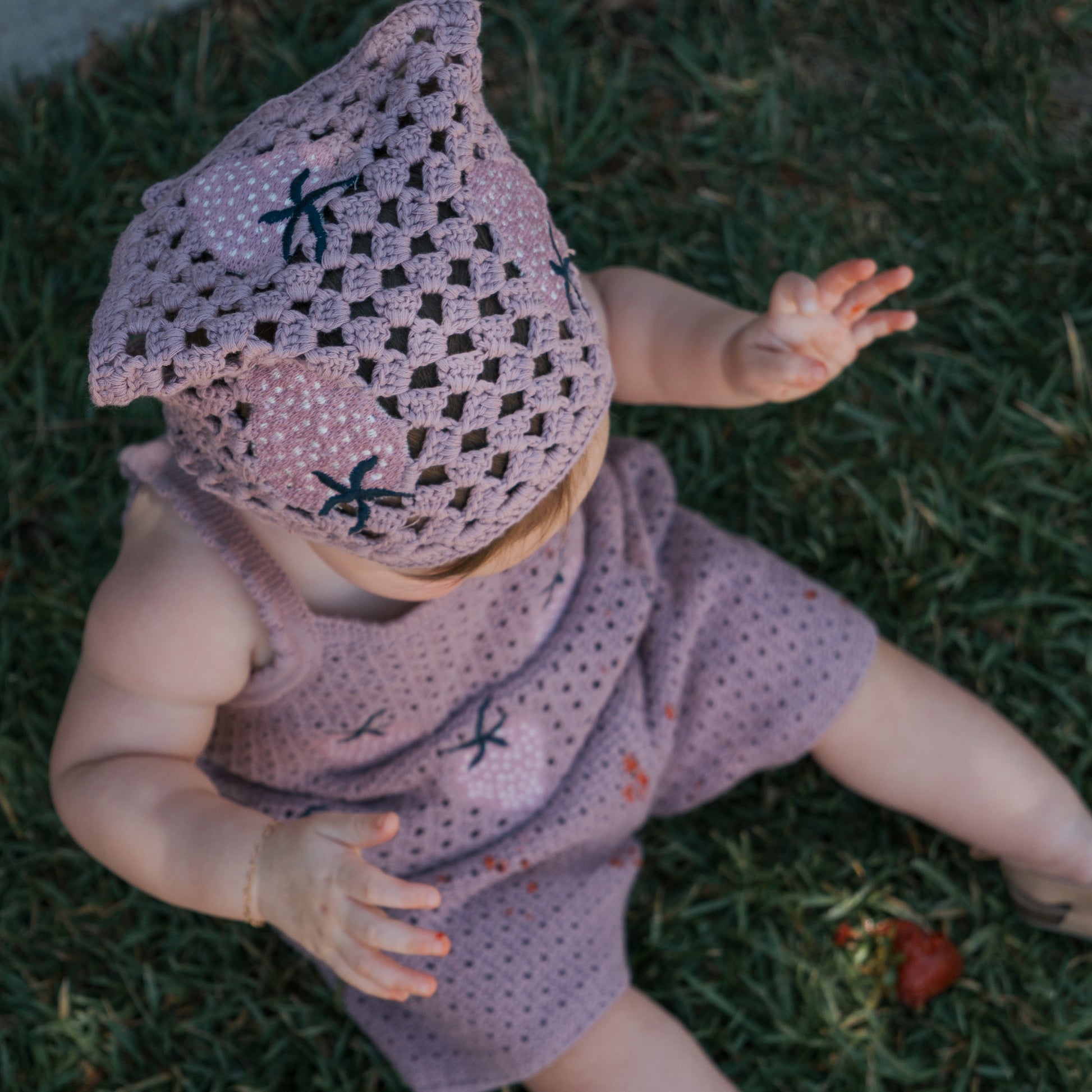 A baby wearing the forever french baby Knit Head Scarf | Matilda sits on grass with arms outstretched, viewed from above, the soft cotton yarn ensuring extra comfort.
