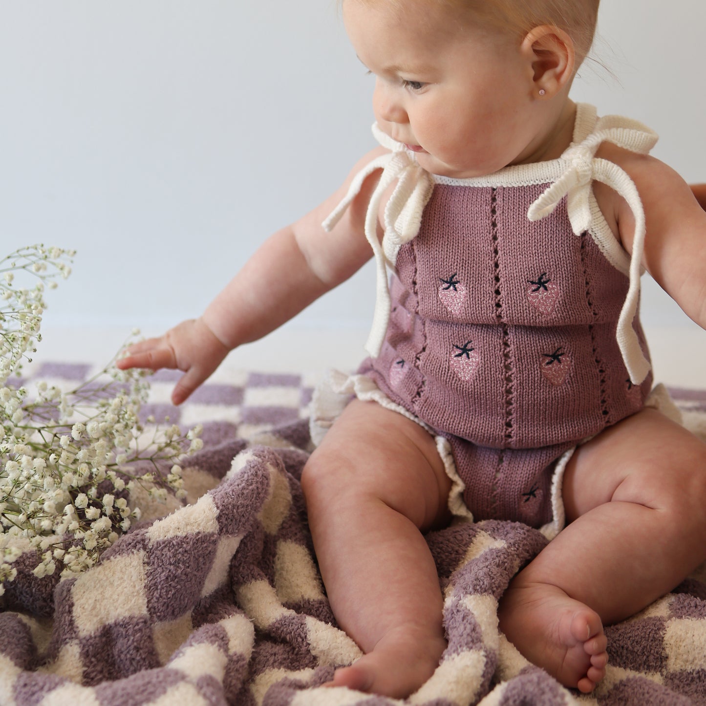 A baby in the forever french baby Knit Ruffle Romper | Matilda with adjustable tie straps and snap closures sits on a purple and white checkered blanket, reaching for a bunch of white baby's breath flowers.