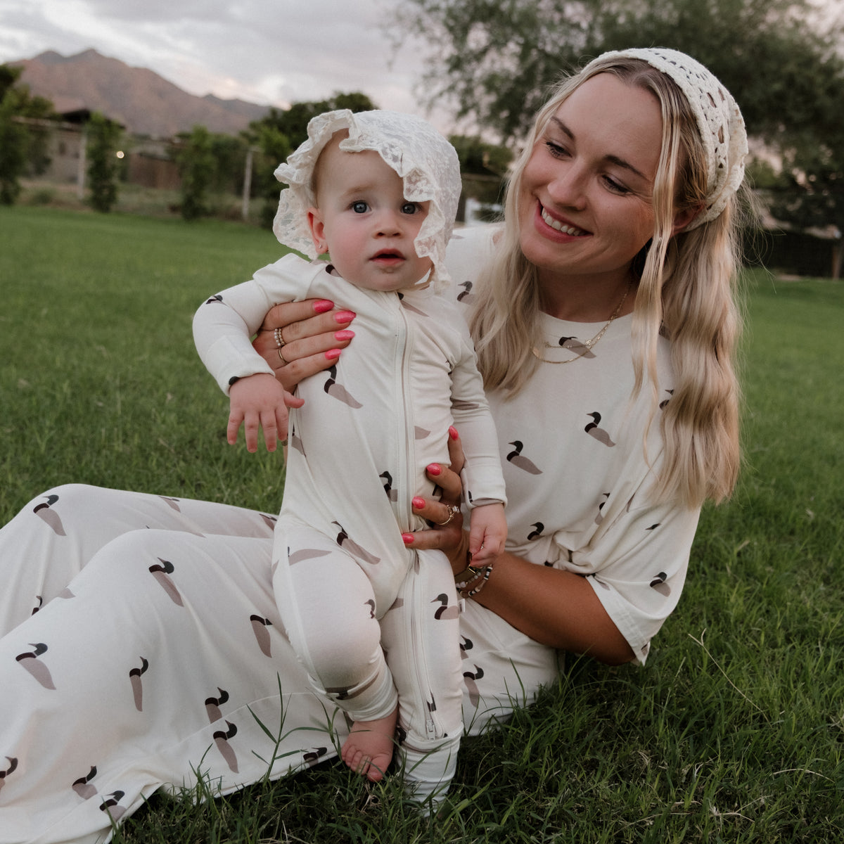 A smiling woman in a patterned dress sits on the grass, holding a baby dressed in forever french baby Bamboo Zip Pajamas | Mallard and a lacy bonnet. Trees, mountains, and a cloudy sky are in the background.