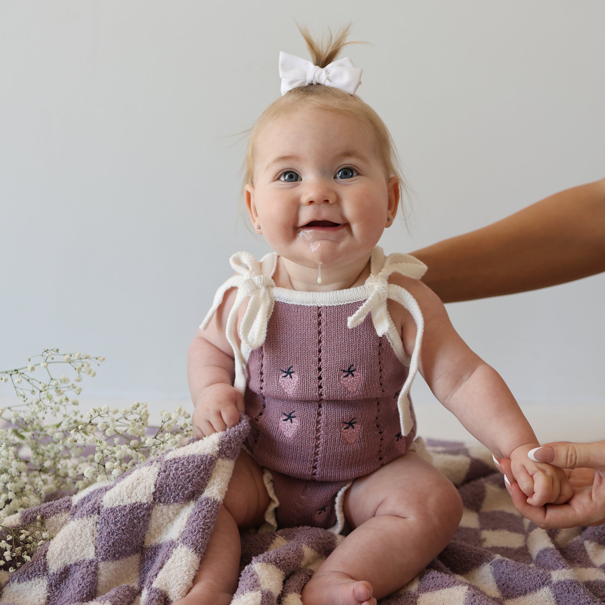A smiling baby with a white bow sits on a purple and white checkered blanket, wearing the forever french baby Knit Ruffle Romper | Matilda with adjustable tie straps. An adult holds her hand as baby's breath flowers rest beside her.