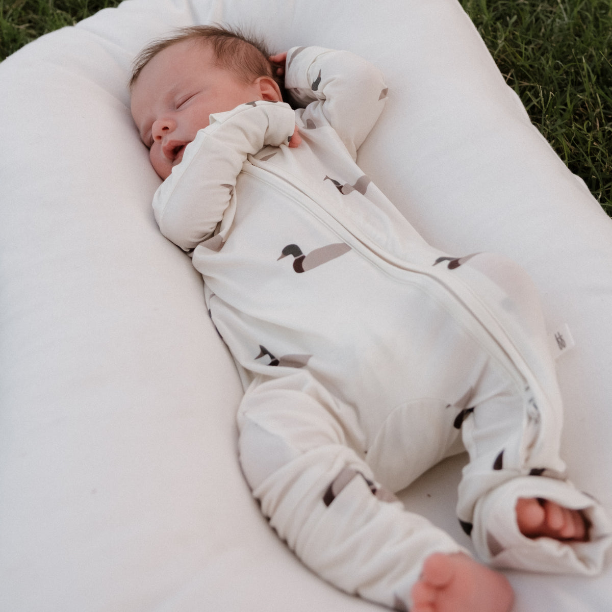 A baby sleeps peacefully on a white cushion outdoors, wearing forever french baby’s Bamboo Zip Pajamas in the Mallard print—a hypoallergenic, white zip-up onesie featuring gray and black patterns.