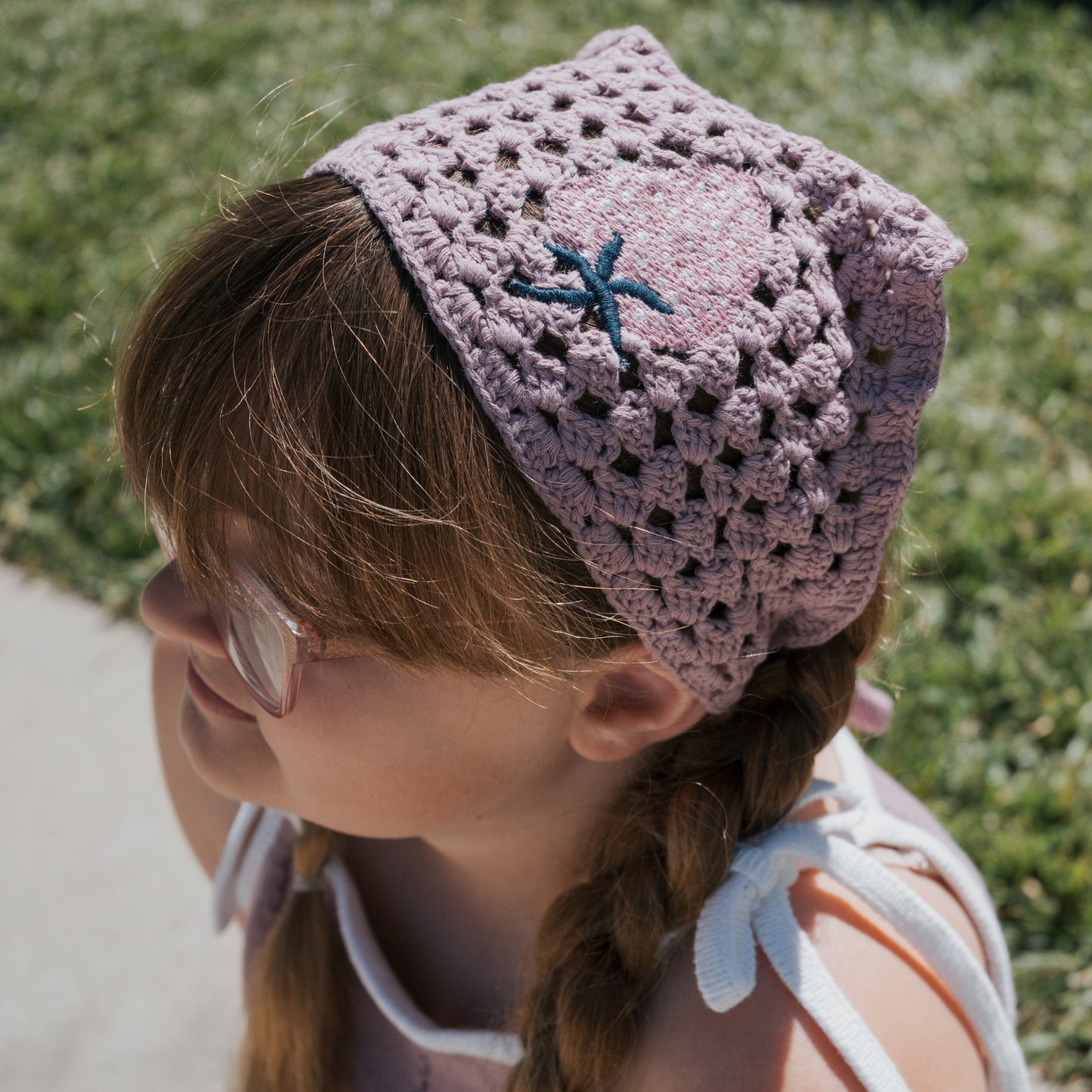 A young girl with brown braided hair wears sunglasses, a pink crocheted headscarf with a blue flower, and the forever french baby Knit Dress | Matilda—a white 100% cotton dress with adjustable tie straps—sitting by grass on a sunny day.