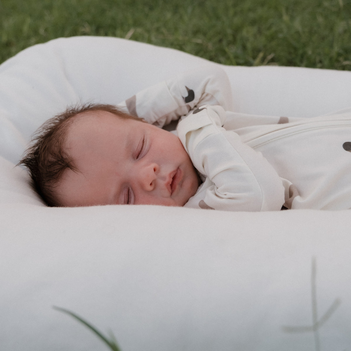 A baby with dark hair sleeps peacefully on a white cushion outdoors, dressed in forever french baby’s Bamboo Zip Pajamas in Mallard, featuring a subtle pattern. Green grass is visible in the background.