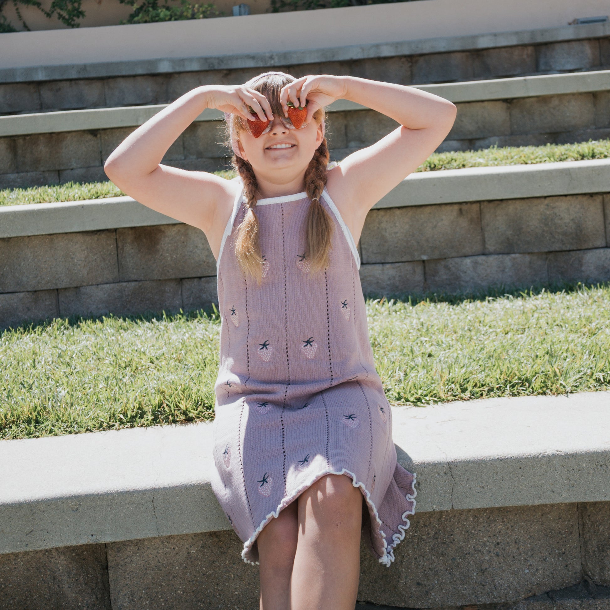 A young girl wears the forever french baby Knit Dress | Matilda with adjustable tie straps as she sits on outdoor steps, playfully holding peach slices over her eyes in the sunlight, with grass and a stone wall in the background.