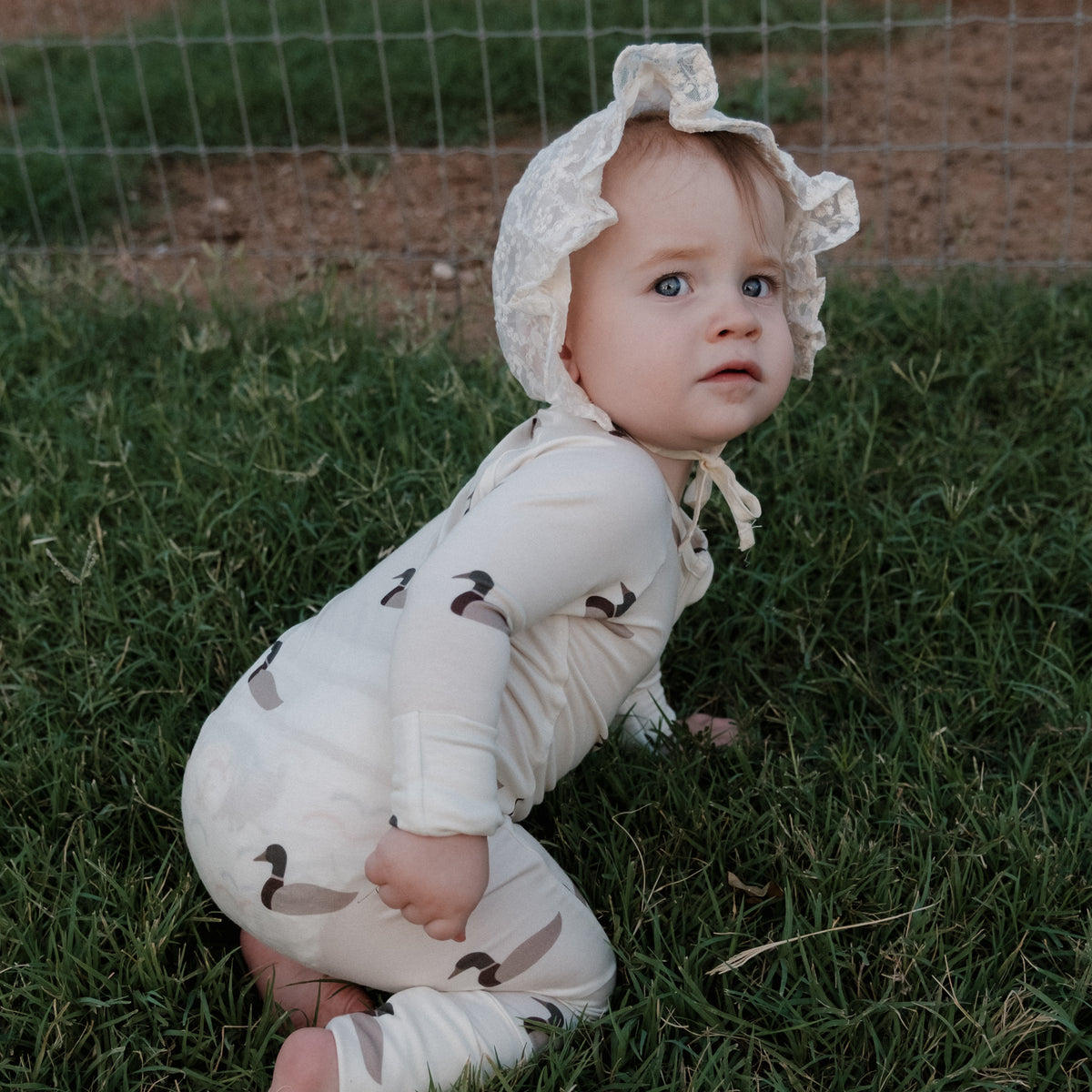 A baby wearing a bonnet and forever french baby Bamboo Zip Pajamas in the Mallard pattern sits on green grass near a wire fence, glancing slightly over their shoulder toward the camera.