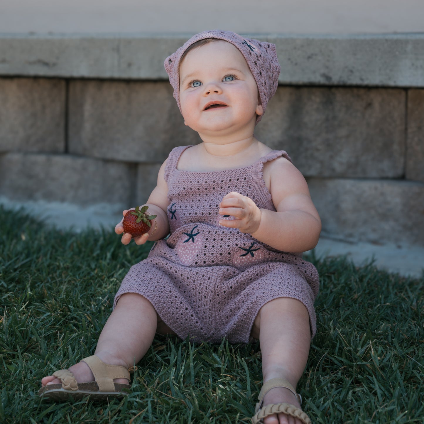 A baby dressed in the Girl's Knit Short Set | Matilda by forever french baby—featuring soft 100% cotton lavender knit fabric and a matching headscarf—sits on the grass holding a strawberry, gazing upward with a stone wall in the background.