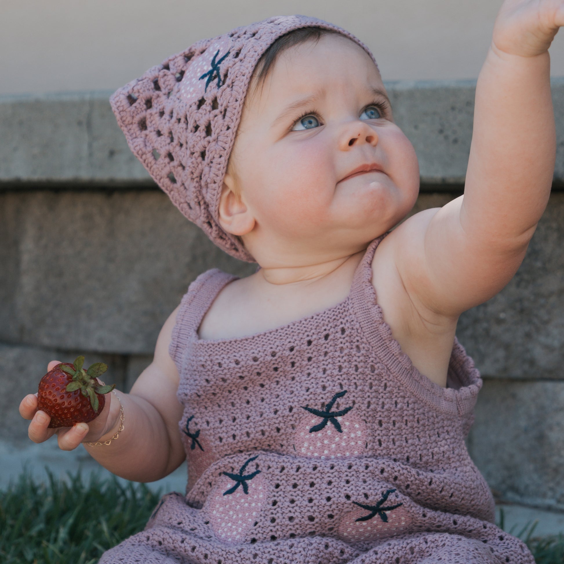 A baby, outdoors and gazing into the distance, wears the "Matilda" Knit Head Scarf by forever french baby—a soft cotton crochet accessory—along with a matching outfit, holding a strawberry in one hand and reaching upward with the other.