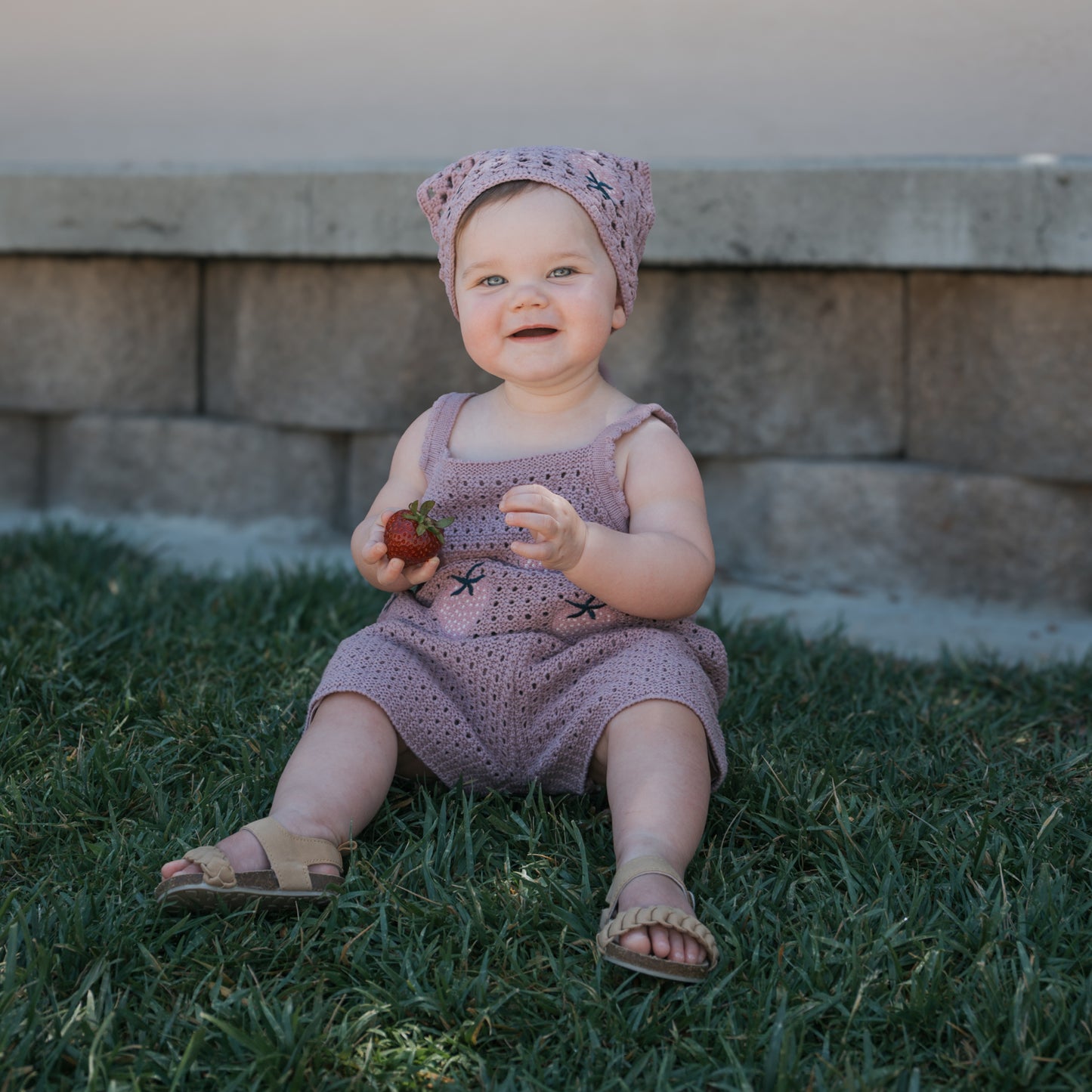 A smiling baby sits on the grass holding a strawberry, wearing the forever french baby Knit Head Scarf | Matilda in soft purple cotton yarn, with a stone wall in the background.