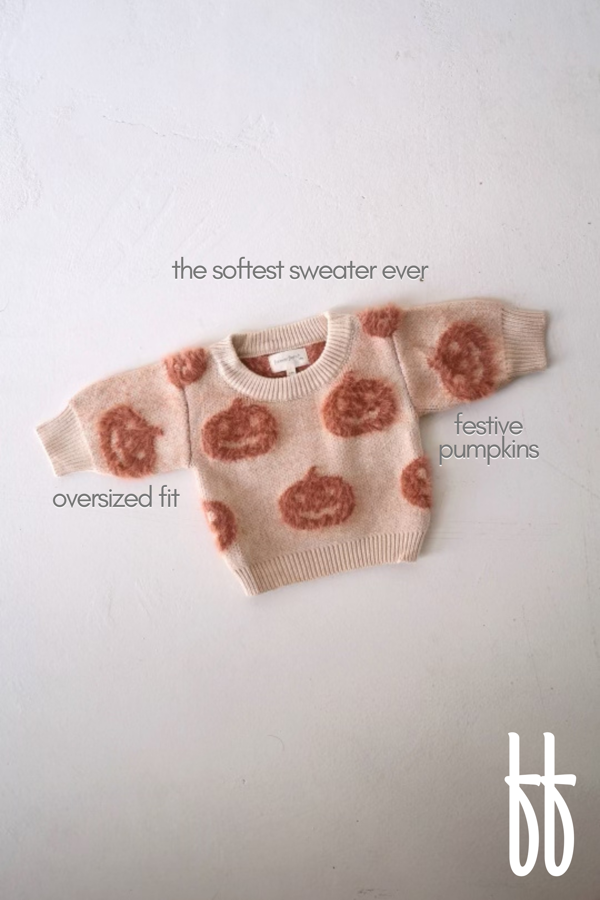 A cream-colored, oversized sweater boasting a soft texture and adorned with orange festive pumpkin designs is laid flat on a white surface. Text on the image labels it as "the ultra-soft Cozy Sweater | Pumpkin" by forever french baby, with highlights on "festive pumpkins" and "oversized fit.