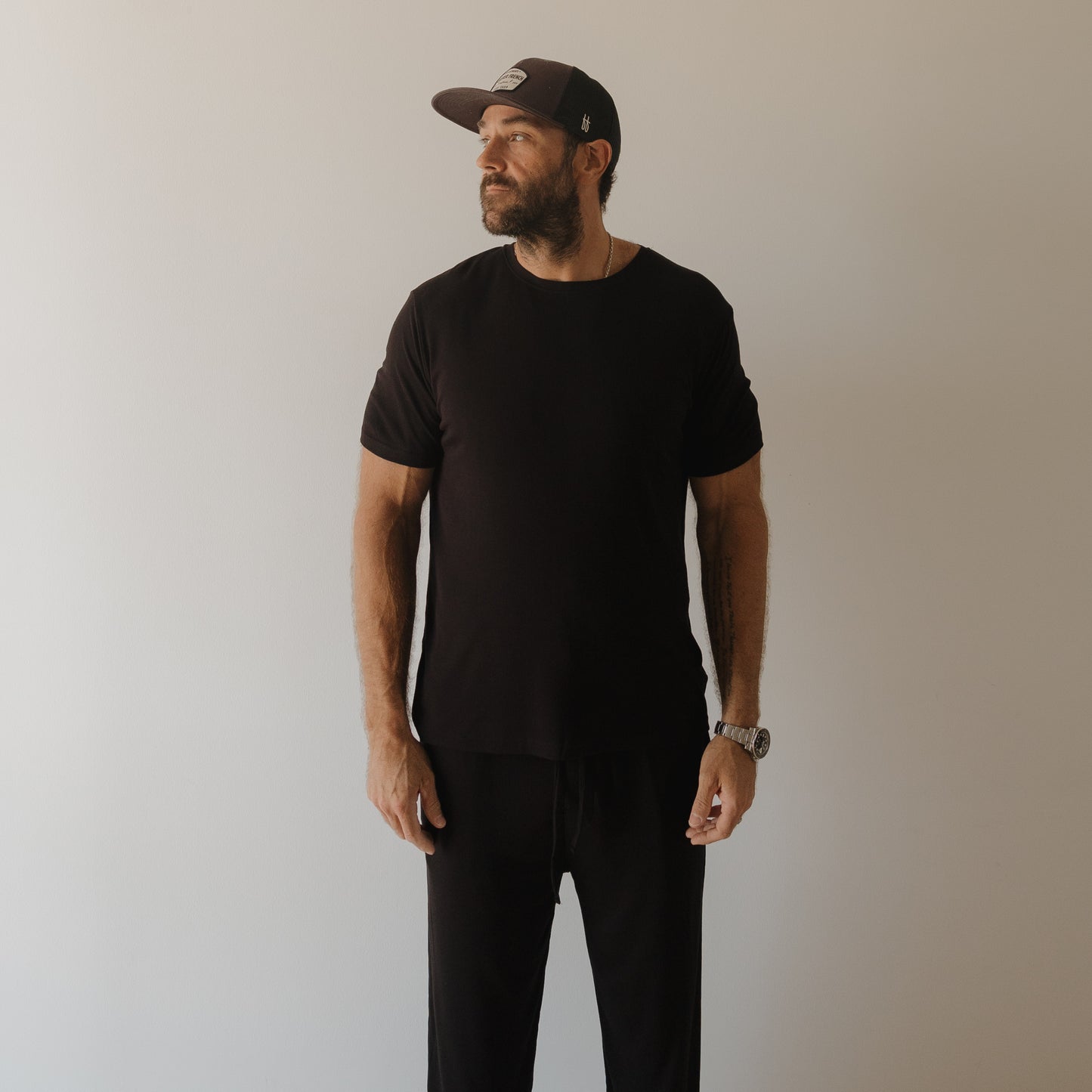 A bearded man wears forever french baby’s Men's Bamboo Short Sleeve Pajama | Ribbed Black, standing against a light wall and gazing left with a neutral expression, his relaxed outfit perfect for comfort.