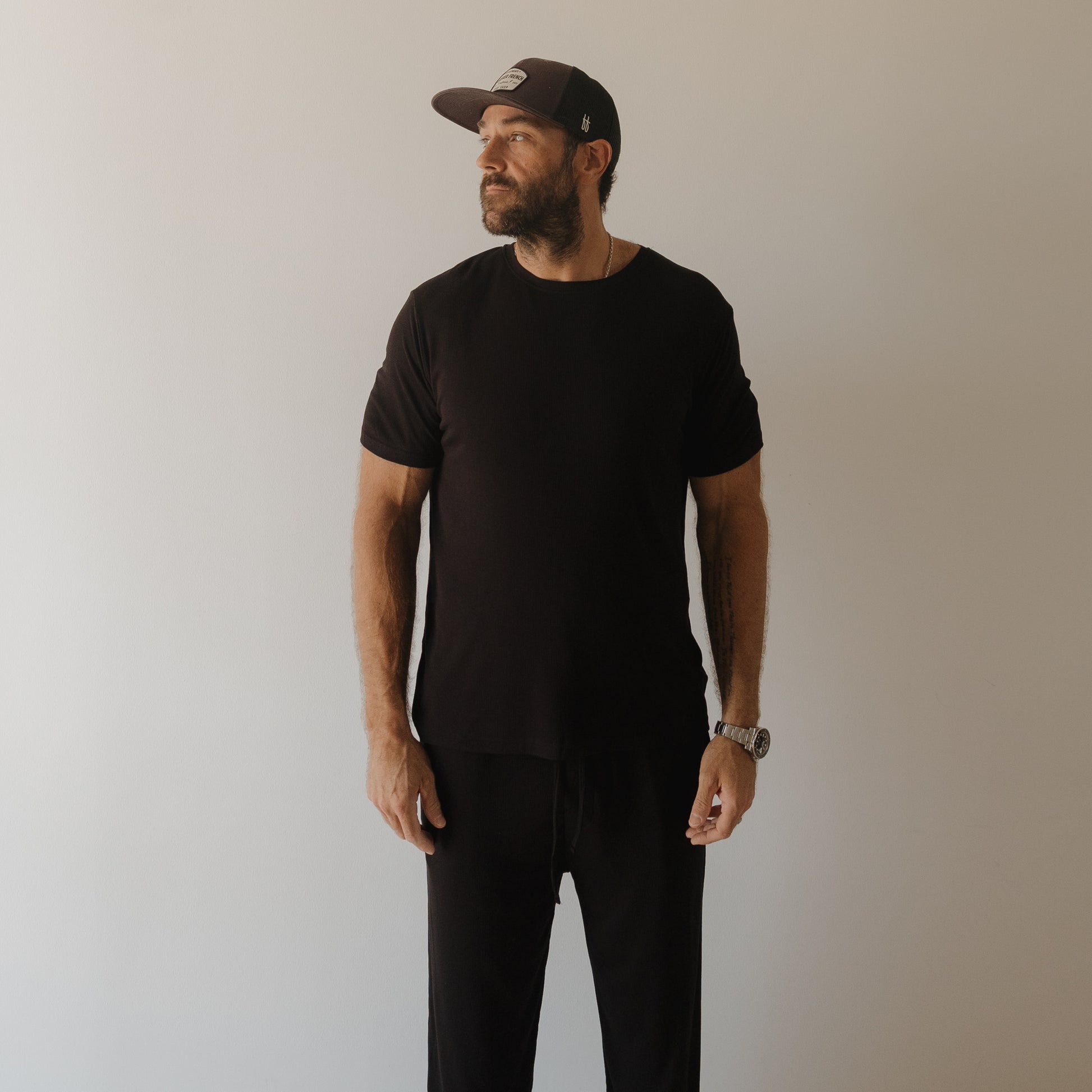 A bearded man wears forever french baby’s Men's Bamboo Short Sleeve Pajama | Ribbed Black, standing against a light wall and gazing left with a neutral expression, his relaxed outfit perfect for comfort.