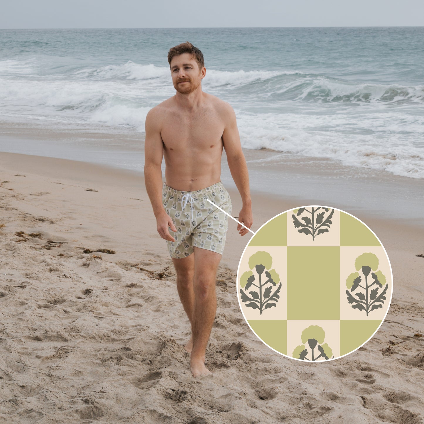 A man strolls a sandy beach in forever french baby Men's Board Short | Marigold Check, featuring cream and green checker print with dark floral motifs. Behind him, waves roll in under a cloudy sky.