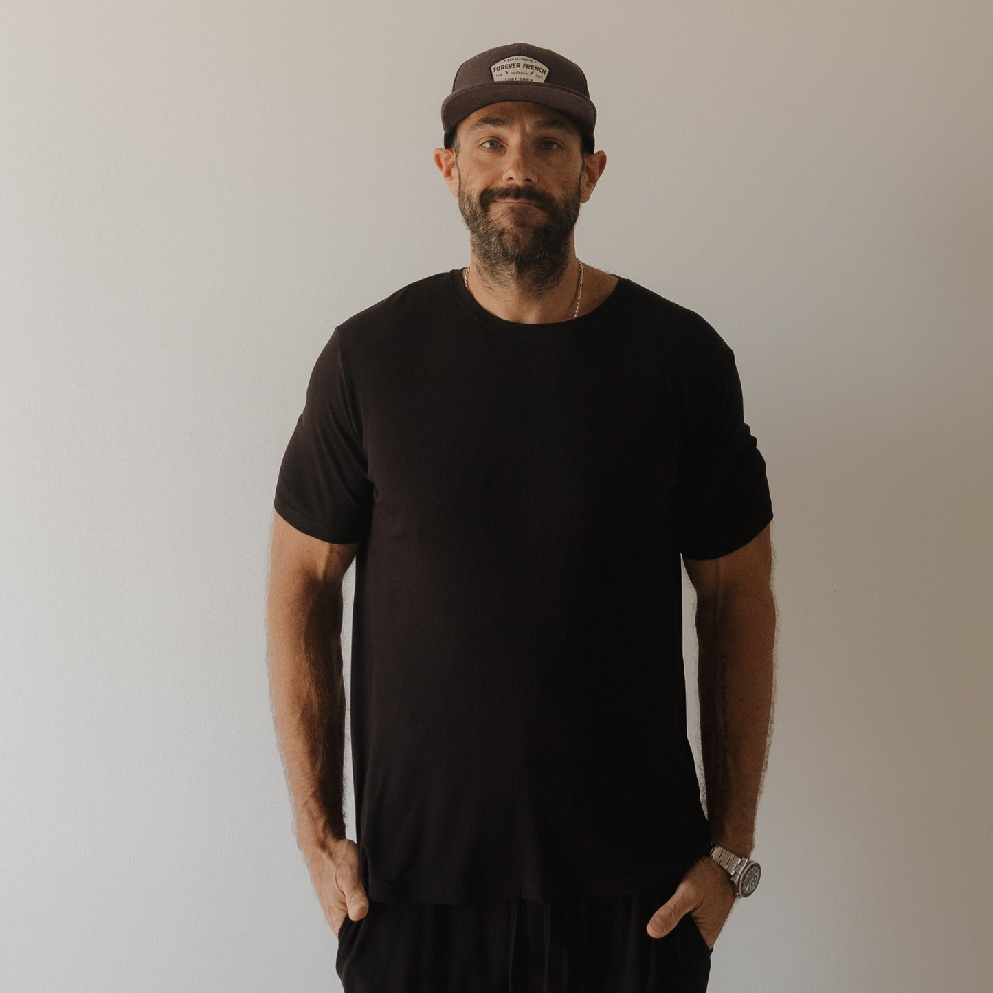 A man in a dark t-shirt, wristwatch, and cap stands against a light wall with hands in pockets—highlighting the comfort of forever french baby Men's Bamboo Short Sleeve Pajama | Ribbed Black.