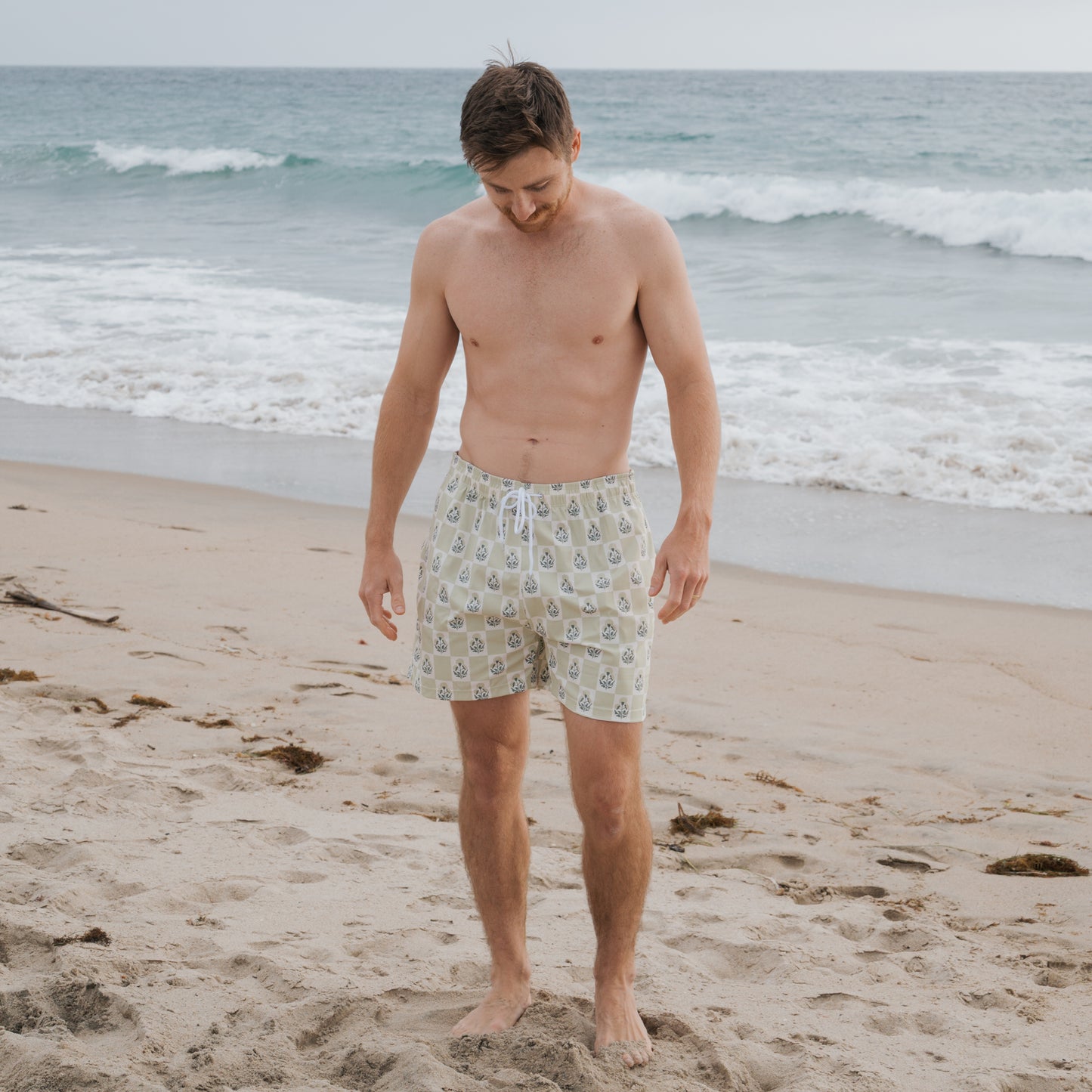 A shirtless man wearing forever french baby Men's Board Short in Marigold Check stands barefoot on the beach, looking down at the sand as gentle waves roll in behind him.