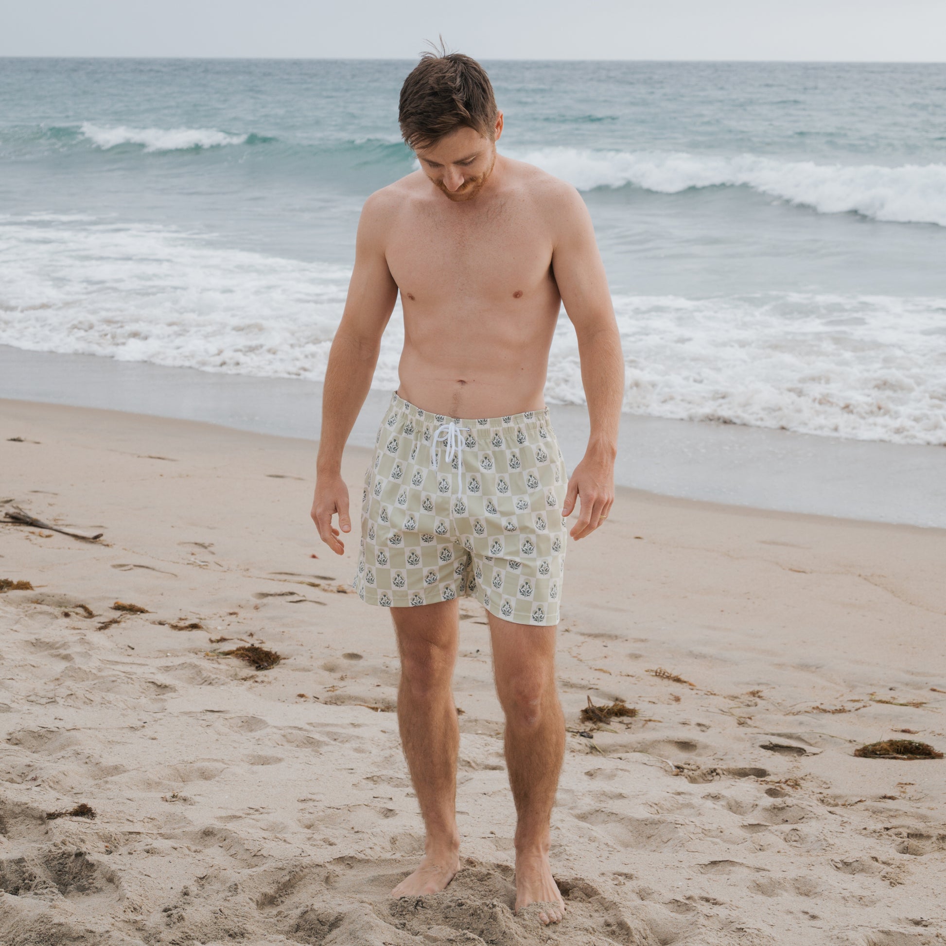 A shirtless man wearing forever french baby Men's Board Short in Marigold Check stands barefoot on the beach, looking down at the sand as gentle waves roll in behind him.