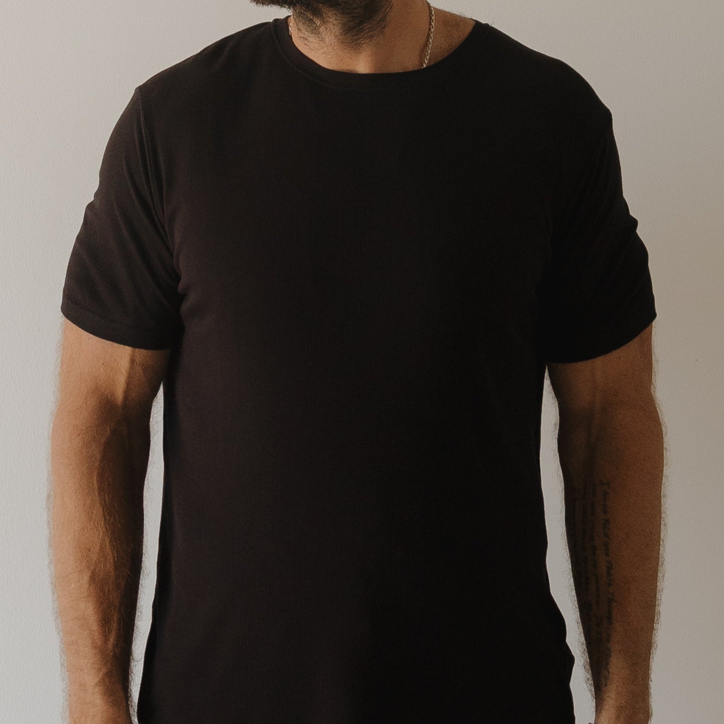 A bearded person wears the forever french baby Men's Bamboo Short Sleeve Pajama in Ribbed Black against a neutral background. Their face is partly obscured, and a forearm tattoo peeks out, showcasing the stylish hypoallergenic sleepwear.