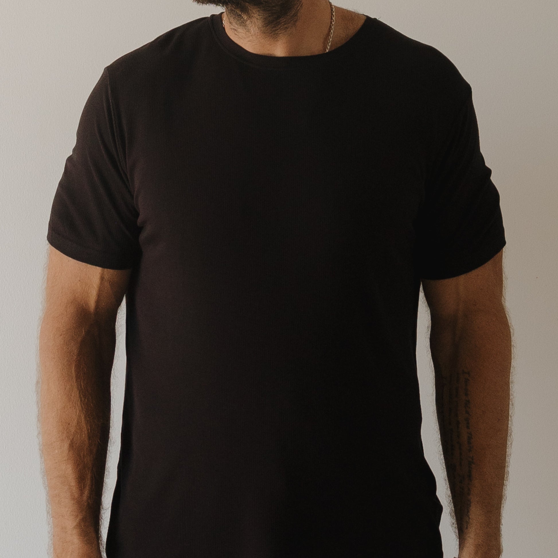 A bearded person wears the forever french baby Men's Bamboo Short Sleeve Pajama in Ribbed Black against a neutral background. Their face is partly obscured, and a forearm tattoo peeks out, showcasing the stylish hypoallergenic sleepwear.