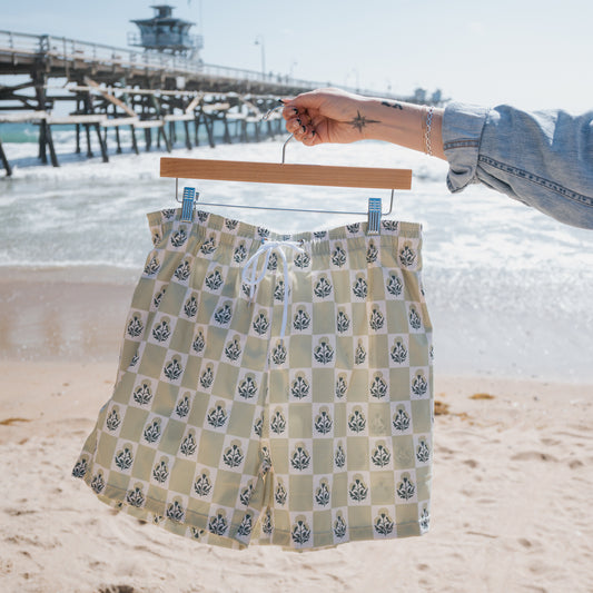 A person holds a forever french baby Men's Board Short | Marigold Check, featuring floral and geometric checker print, in front of a sandy beach, ocean, pier, and blue sky.