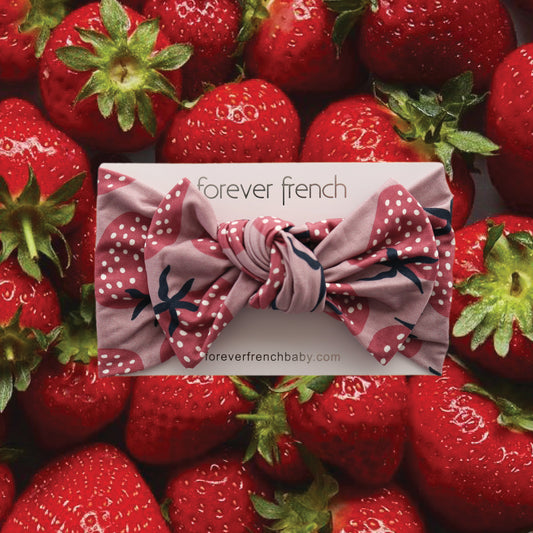 A Bamboo Head Wrap | Matilda by forever french baby, featuring a pink strawberry-themed design, is shown against fresh strawberries. The brand name appears on the product’s packaging, highlighting this adorable bamboo headband.