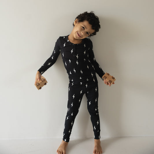 A young child with curly hair smiles barefoot against a white wall, holding wooden toy cars and wearing forever french baby Bamboo Two Piece Pajamas | Midnight Lightning Bolt—soft, hypo-allergenic sleepwear perfect for sensitive skin.