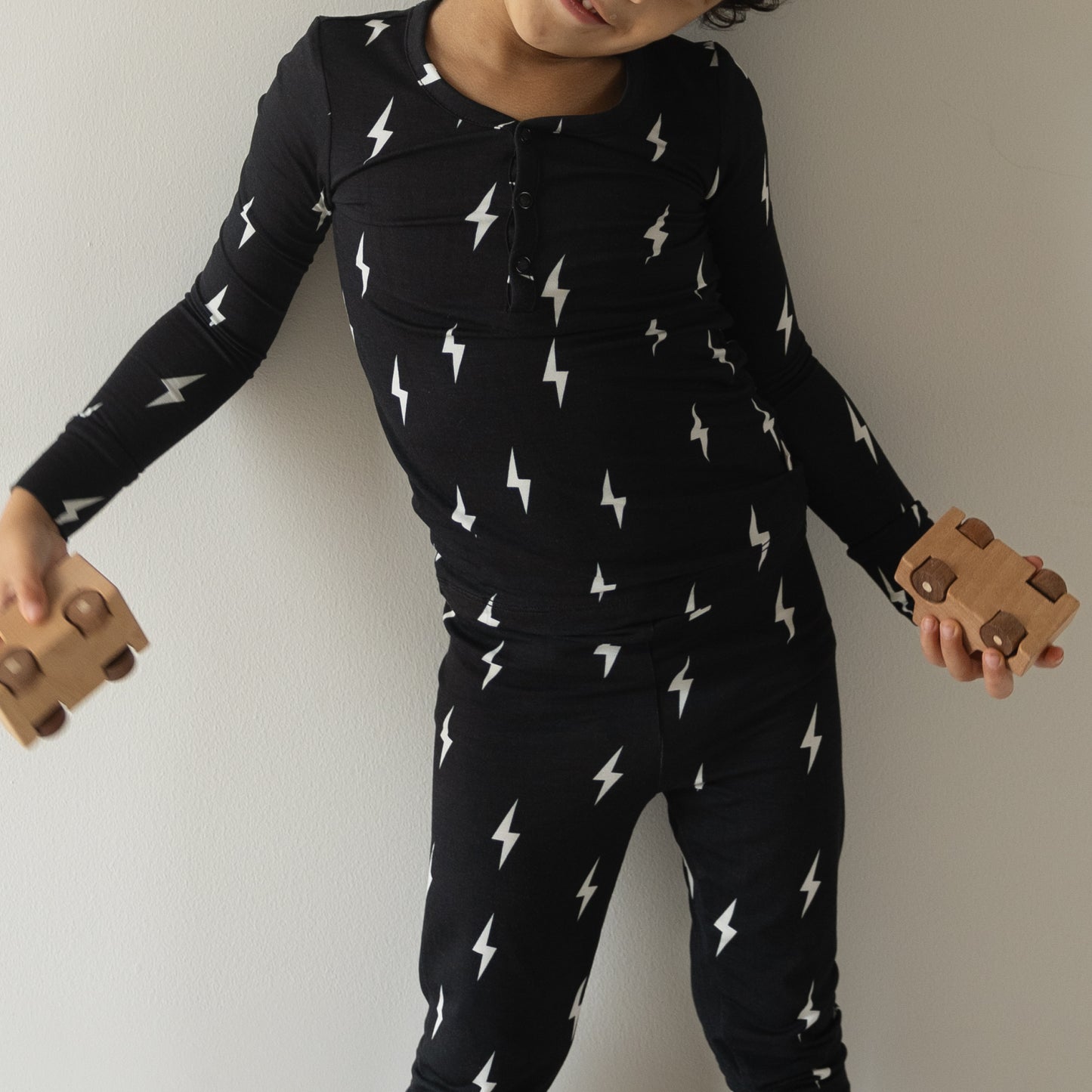 A child in forever french baby Bamboo Two Piece Pajamas | Midnight Lightning Bolt holds two wooden toy cars while standing against a plain white wall.