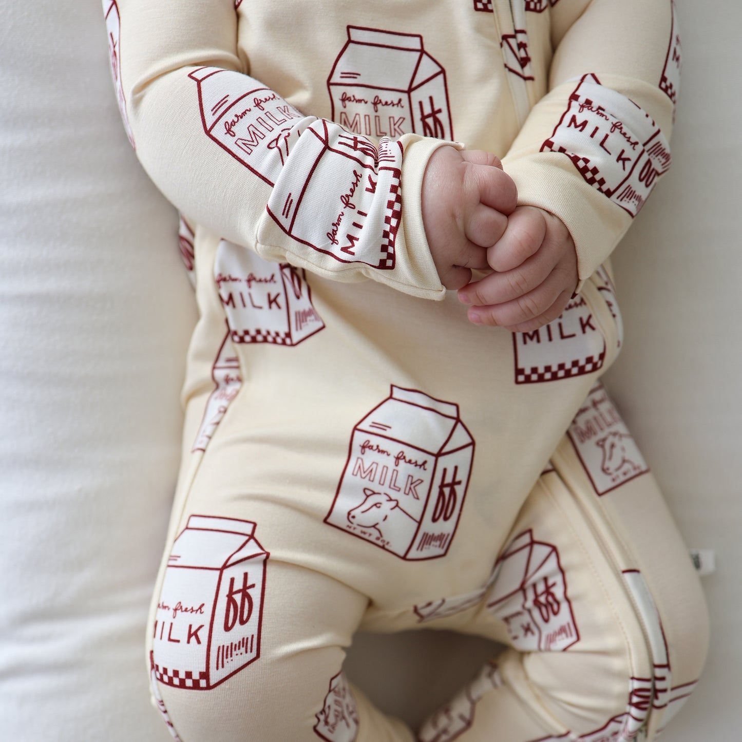 Wearing forever french baby's Bamboo Zip Pajamas | Milk—a cream onesie with red milk carton prints—a baby sits on a white surface, hands clasped, with only their lower face and body visible.