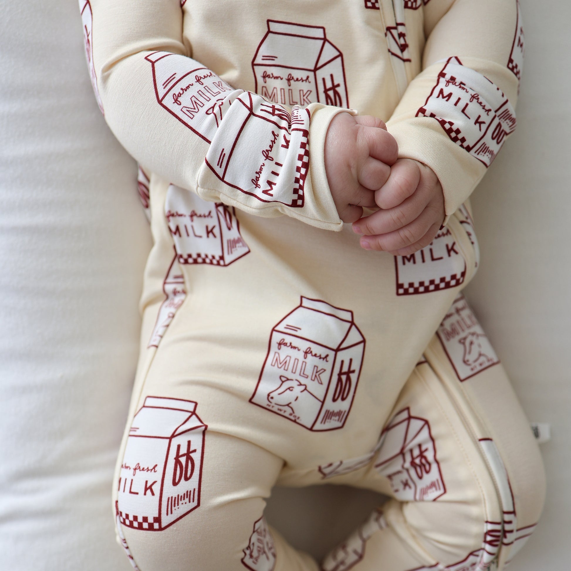 Wearing forever french baby's Bamboo Zip Pajamas | Milk—a cream onesie with red milk carton prints—a baby sits on a white surface, hands clasped, with only their lower face and body visible.