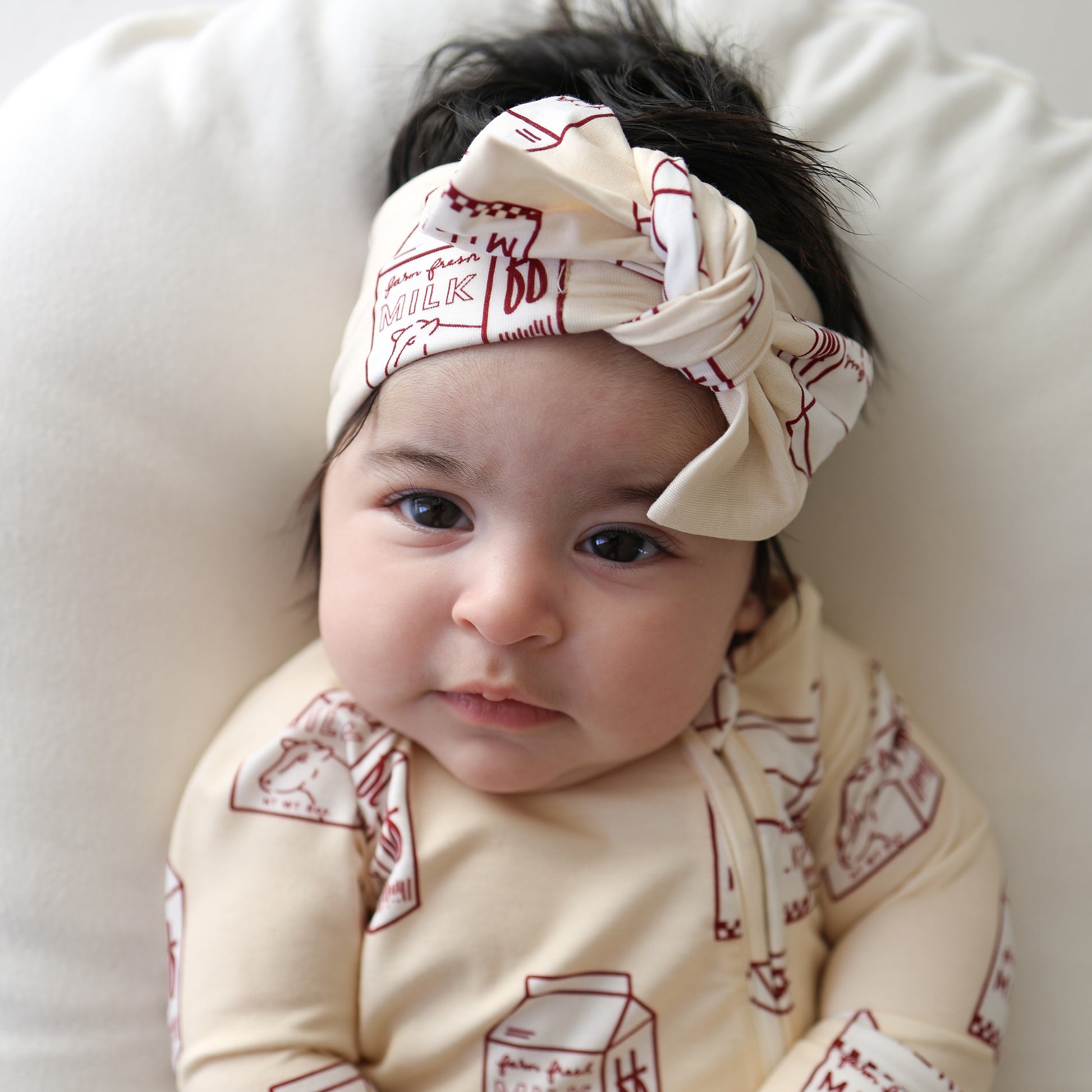 A baby in a cream forever french baby outfit and a Bamboo Head Wrap | Milk with red milk carton prints lies on a white cushion, gazing calmly at the camera.