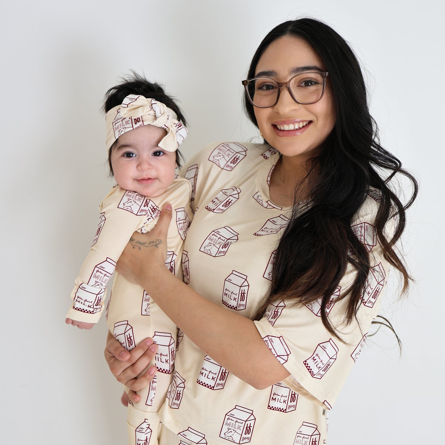 A smiling woman with long dark hair and glasses holds a happy baby. Both wear matching forever french baby Bamboo Zip Pajamas | Milk, featuring cream hypoallergenic fabric with red milk carton prints. The baby also has a matching headband on a plain white background.