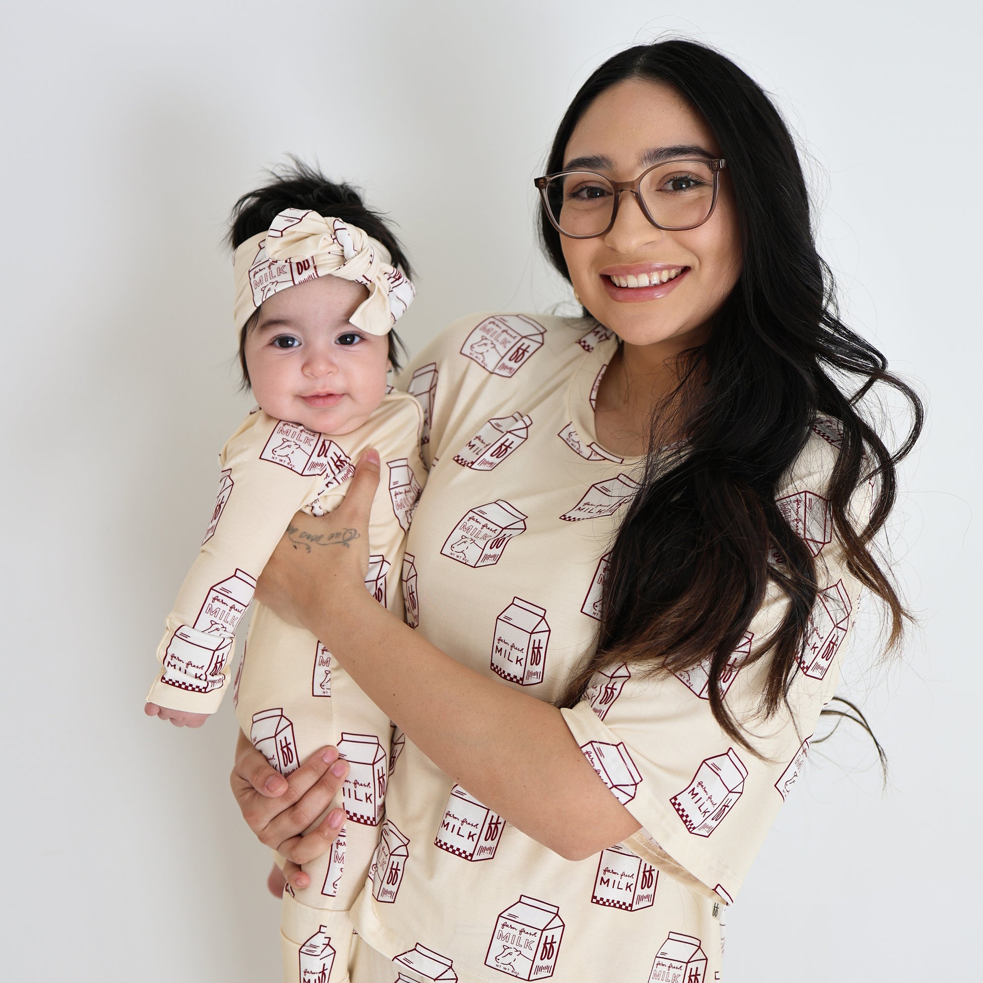 A smiling woman with long dark hair and glasses holds a happy baby. Both wear matching forever french baby Bamboo Zip Pajamas | Milk, featuring cream hypoallergenic fabric with red milk carton prints. The baby also has a matching headband on a plain white background.