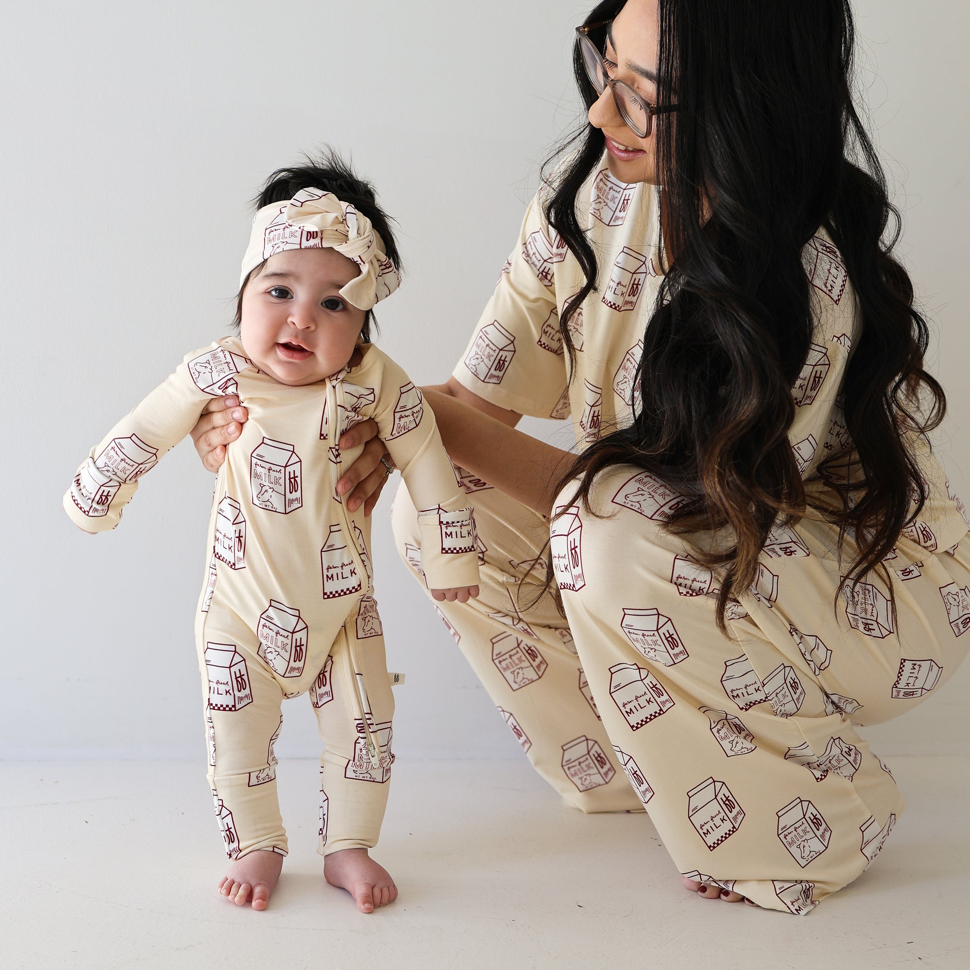 A smiling baby and a woman crouch together in matching forever french baby Bamboo Zip Pajamas | Milk, featuring cute milk carton prints. The hypoallergenic headband and breathable sleepwear offer cozy, stylish comfort.
