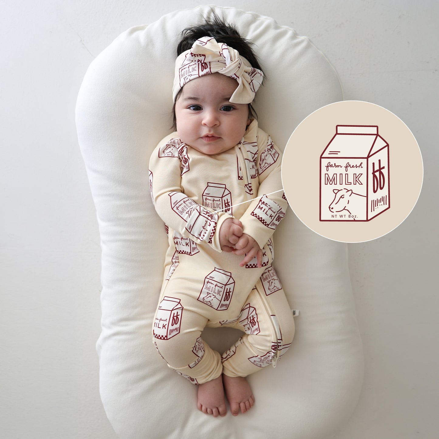 A baby lies on a white cushion, wearing forever french baby's Bamboo Zip Pajamas | Milk and a matching milk-carton headband. An illustrated milk carton sits beside the baby.