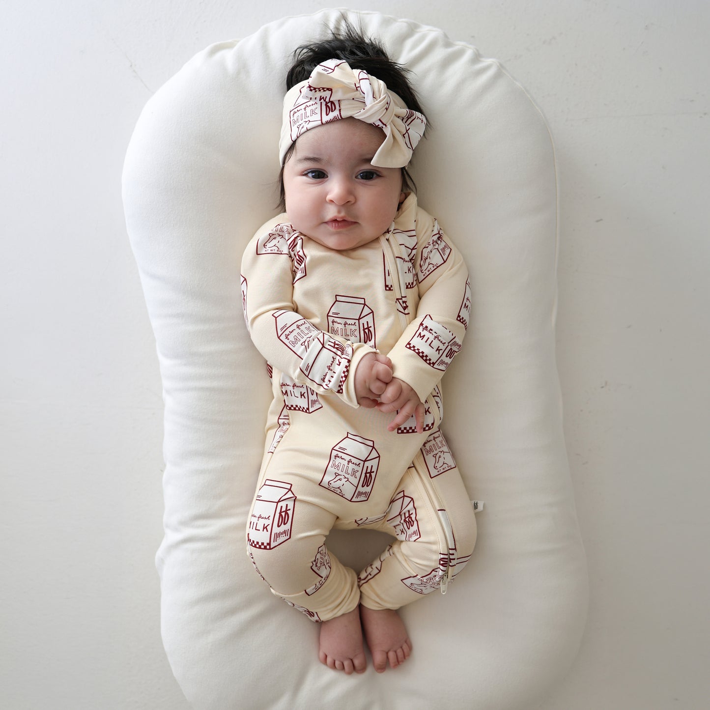 A baby rests on a white cushion, dressed in forever french baby's Bamboo Zip Pajamas | Milk—a cream sleepwear set with red milk carton prints and matching headband—hands folded, feet bare, gazing upward against a light backdrop.