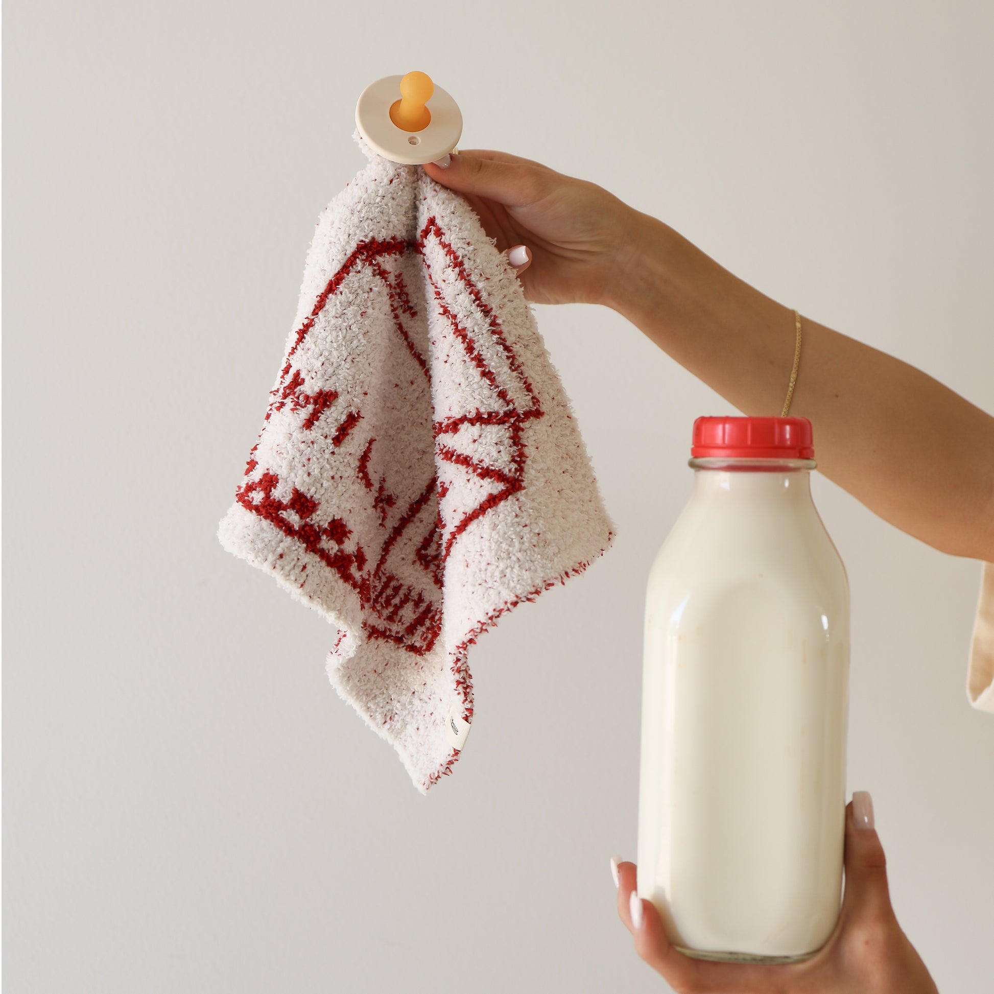A person holds the forever french baby Lovey | Milk microfiber baby blanket and a glass bottle of milk, both set against a plain white background.