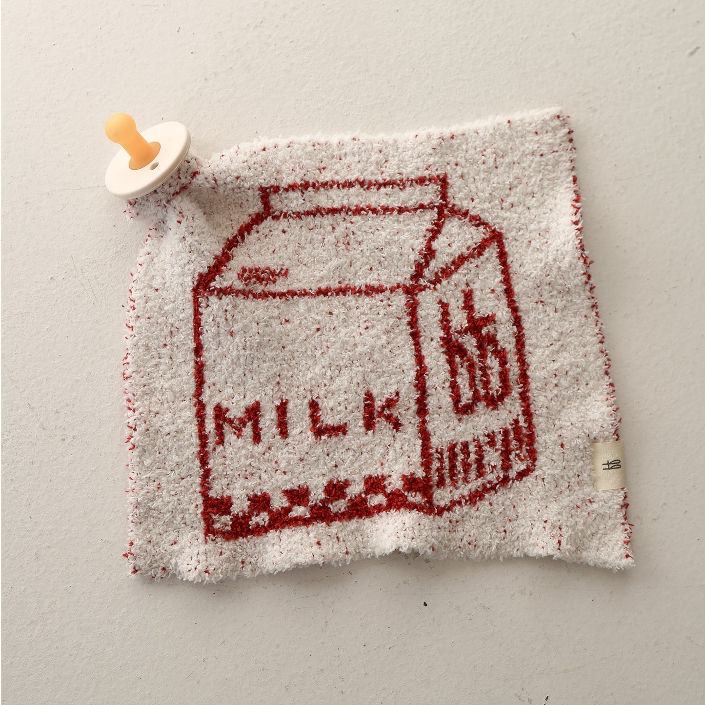 The Lovey | Milk by forever french baby is a white and red textured microfiber blanket with a red milk carton and "MILK" text, plus a pacifier attached to one corner for added practicality and charm.