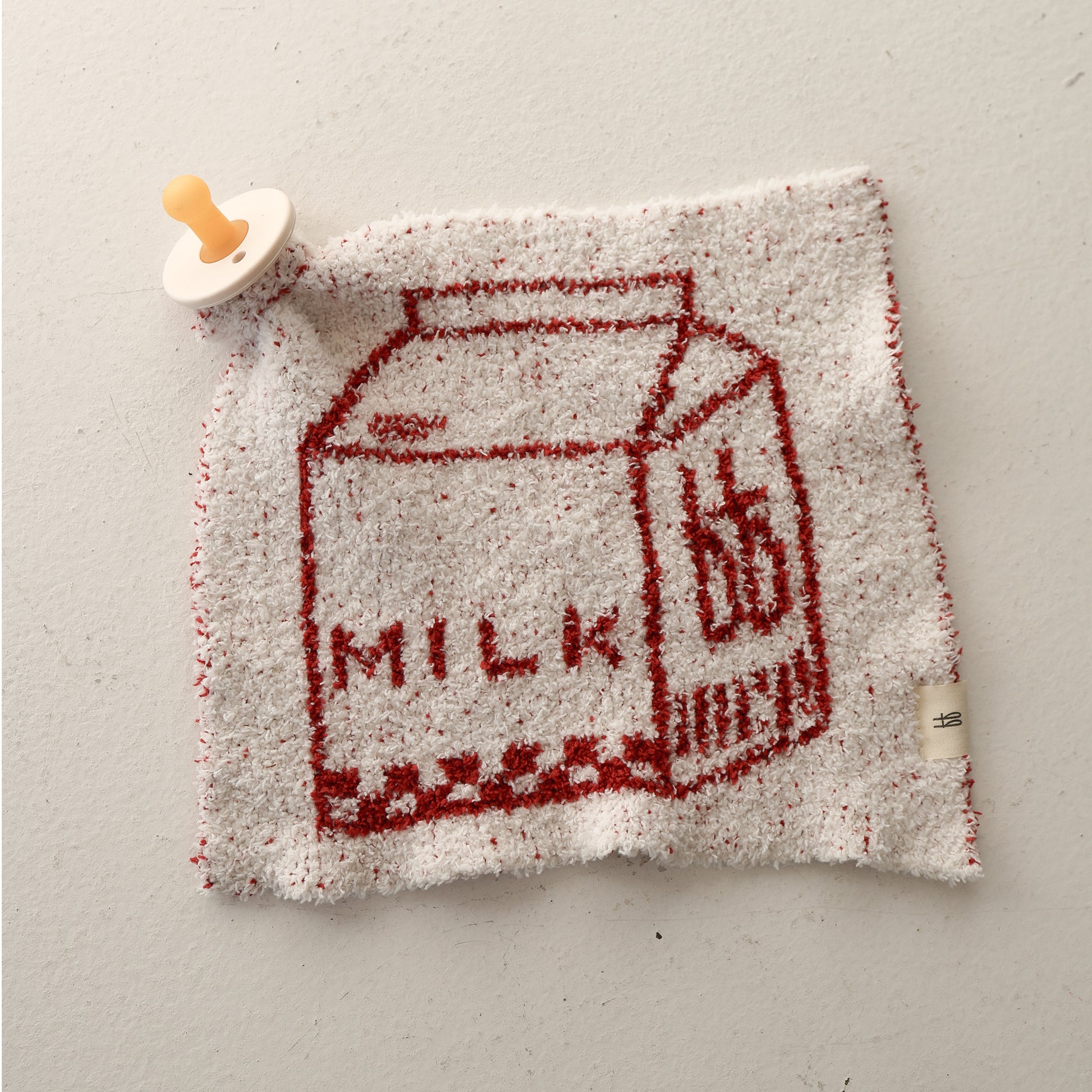The Lovey | Milk by forever french baby is a white and red textured microfiber blanket with a red milk carton and "MILK" text, plus a pacifier attached to one corner for added practicality and charm.