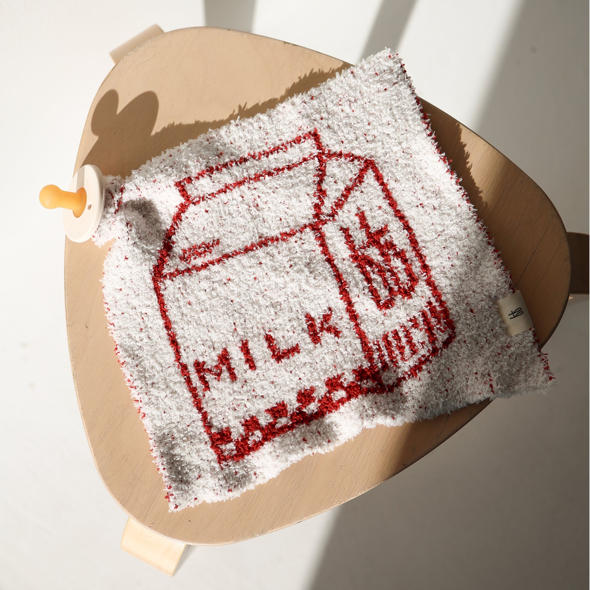 The Lovey | Milk by forever french baby, a white washcloth with a red milk carton and "MILK" design, rests on a light wooden chair beside a pacifier. Sunlight casts gentle shadows, evoking the warmth of a cherished lovey.