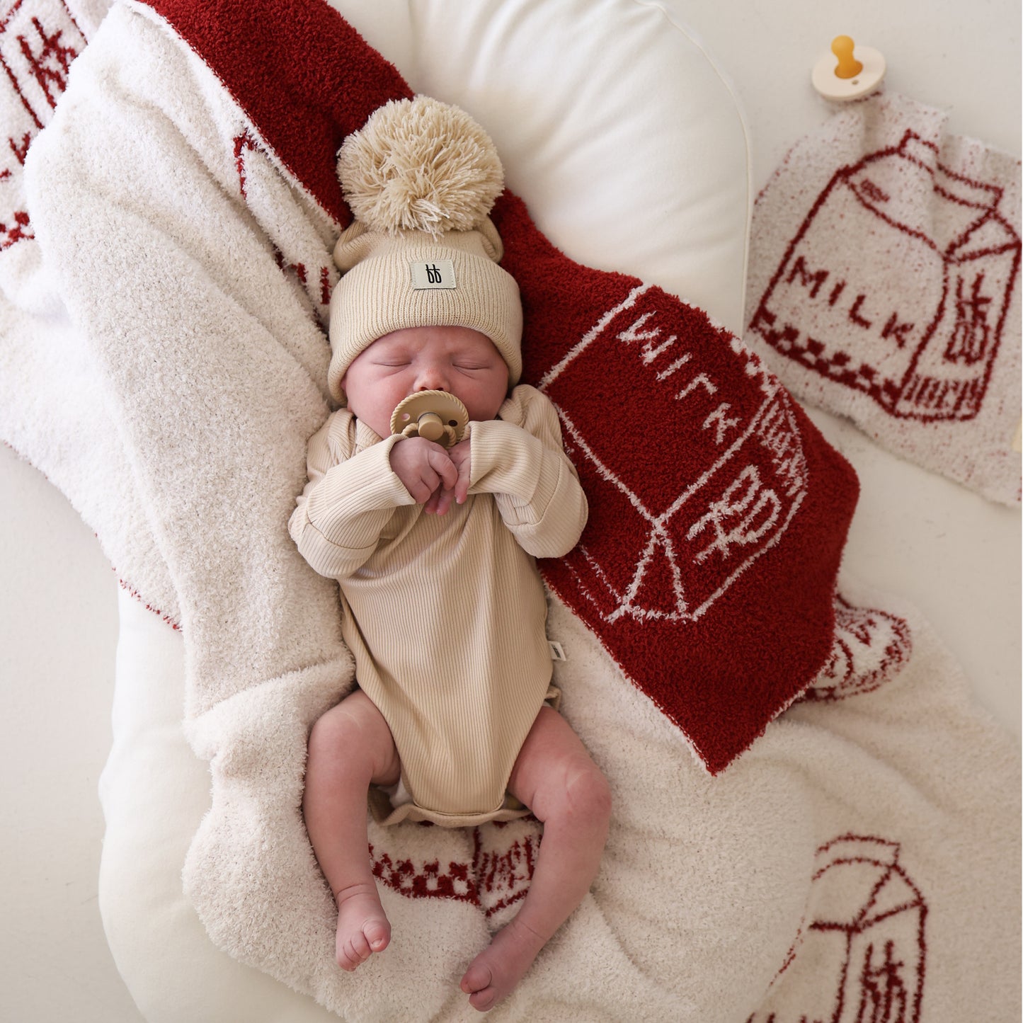 A baby in a beige outfit and pom-pom hat lies on a soft cream microfiber blanket with a pacifier, next to the forever french baby Lovey | Milk—a red and white towel featuring milk carton sketches and the word "milk.