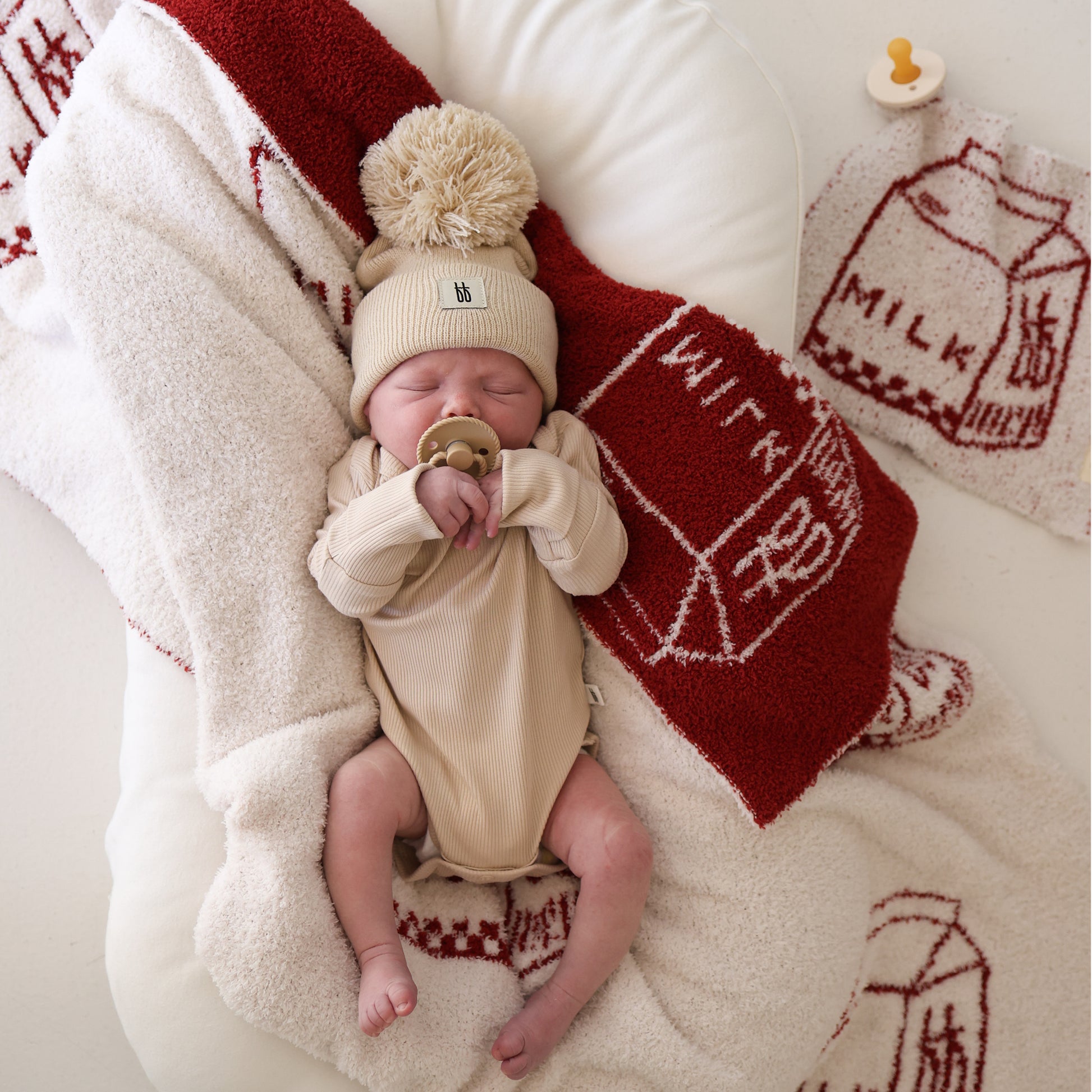 A baby in a beige outfit and pom-pom hat lies on a soft cream microfiber blanket with a pacifier, next to the forever french baby Lovey | Milk—a red and white towel featuring milk carton sketches and the word "milk.